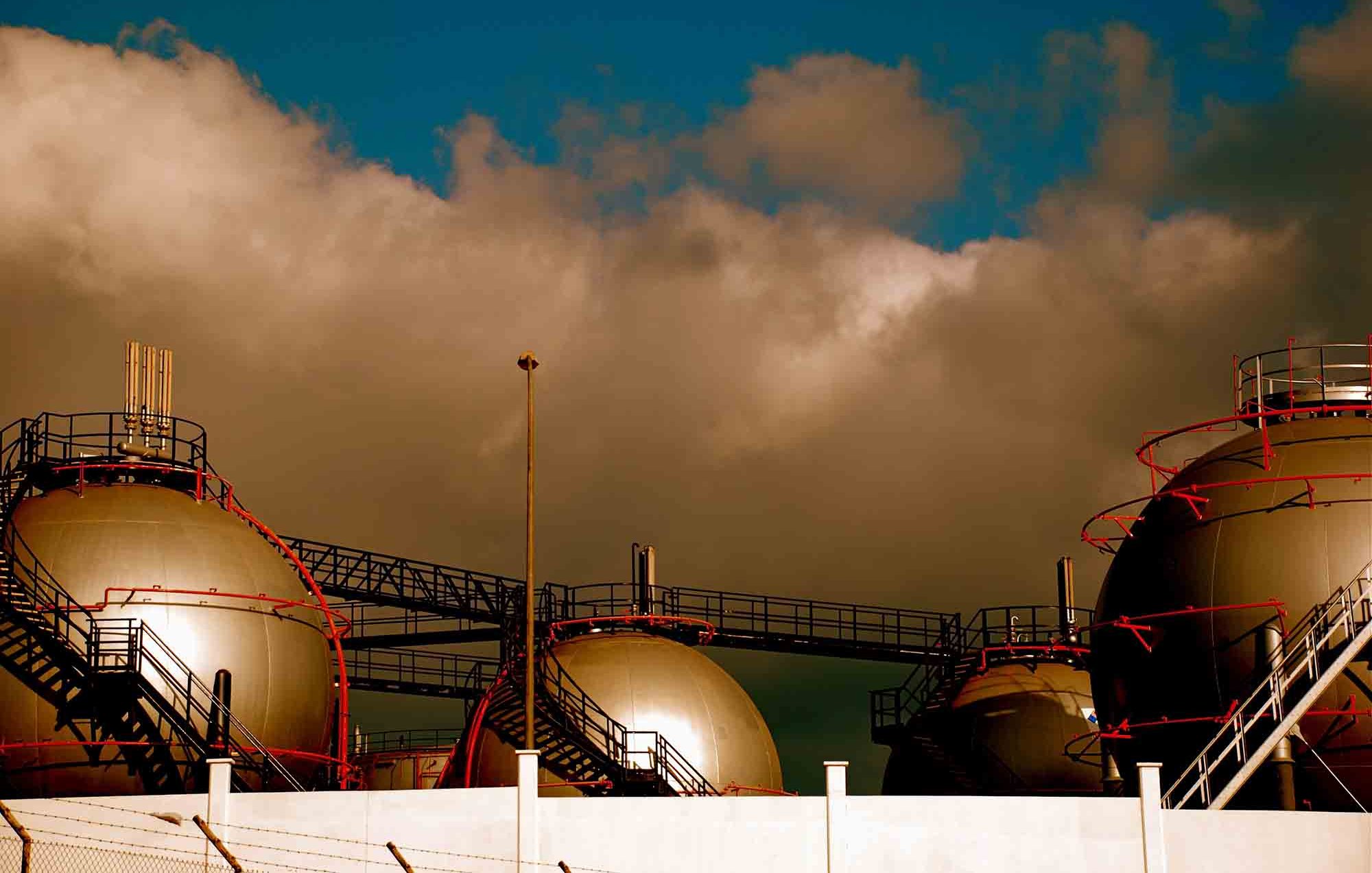 این تصویر نمایی از یک تأسیسات صنعتی را نشان میدهد که شامل چند مخزن بزرگ به شکل کروی است. مخازن روی سکویی قرار دارند و برخی از آنها داکتها و لولههای پیچیدهای دارند که به آنها متصل شدهاند. آسمان در پسزمینه کمی ابری است و رنگها به طور کلی گرم و متنوع به نظر میرسند. اجزای صنعت و بافتهای مختلف تأسیسات به خوبی در تصویر دیده میشوند.