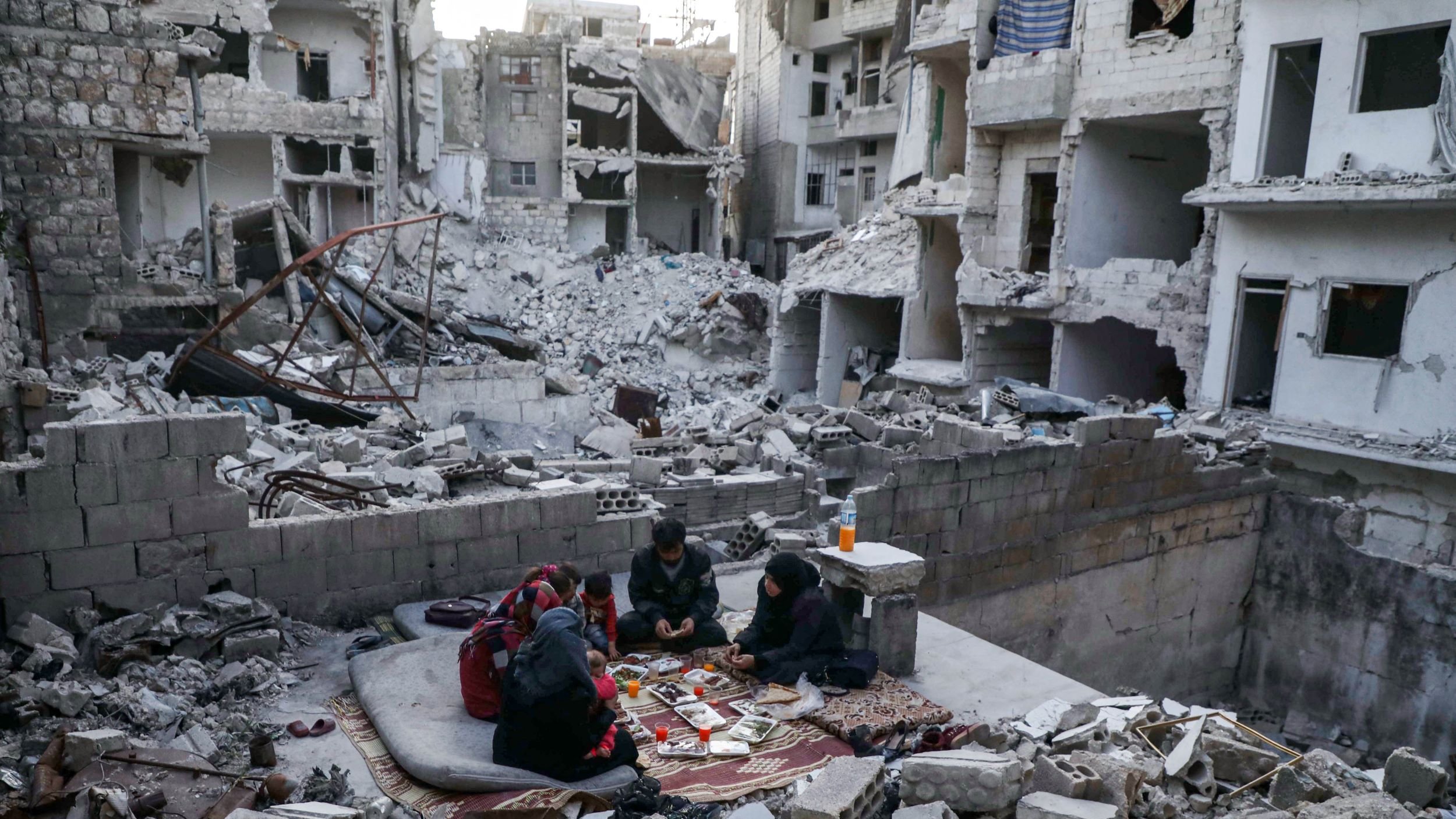 تصویر نشاندهندهی یک منطقه ویران در یک شهر است که آثار تخریب شدید در آن مشهود است. ساختمانها به شدت آسیب دیده و دیوارها و سقفها خراب شدهاند. در وسط این ویرانی، گروهی از افراد نشستهاند و روی زمین، میان خرابهها، در حال بازی یا سرگرمی هستند. فضای کلی تصویر حس ناامیدی و خرابی را منتقل میکند، اما در عین حال حضور انسانها نشانهای از امید و زندگی در شرایط سخت است.