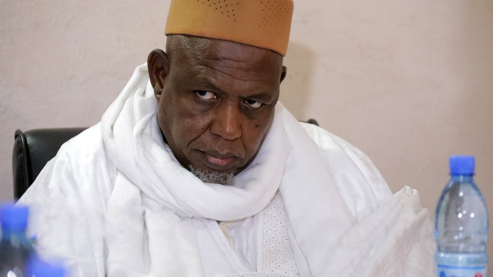 این تصویر یک مرد را نشان میدهد که لباسی سنتی به تن دارد و یک سرپوش قهوهای رنگ بر سر گذاشته است. او در حال نشسته و به جلو خیره شده است، به نظر میرسد که در حال تفکر یا گوش دادن به صحبتهای دیگران است. پسزمینه تصویری ساده و بدون هیچ جزئیات خاصی دارد.