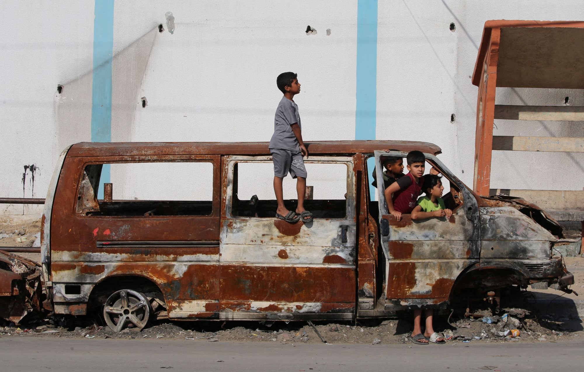 در این تصویر، یک خودرو قدیمی و زنگزده دیده میشود که در یک محیط شهری و آشفته قرار دارد. چند کودک در اطراف این خودرو بازی میکنند و یک کودک بر روی سقف آن ایستاده است. مناظر پشت سر آنها شامل دیوارهای آسیبدیده و ناپایداریهای دیگر است که نشاندهنده شرایط سخت و فرسوده منطقه است. این تصویر احساساتی از کودکی و بازی در شرایط نامناسب را منتقل میکند.