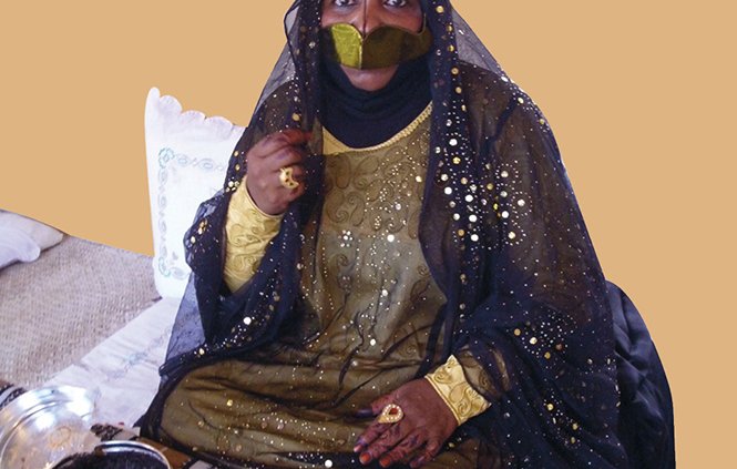 این تصویر یک زن را نشان میدهد که در لباس و پوشش سنتی خود نشسته است. او چهرهاش را با نوار مشکی پوشانده و چشمهایش نمایان است. لباس او شامل رنگهای تیره و زینتهای طلایی است که درخشندگی خاصی دارد. زن در حال نشستن بر روی یک سجاده یا پارچه با خطوط افقی است و در کنار او چند وسیله مانند ظرفهای کوچک دیده میشود. زمینه تصویر نیز به رنگ قهوهای ملایم است و به عکس حس گرم و جذابی میدهد.