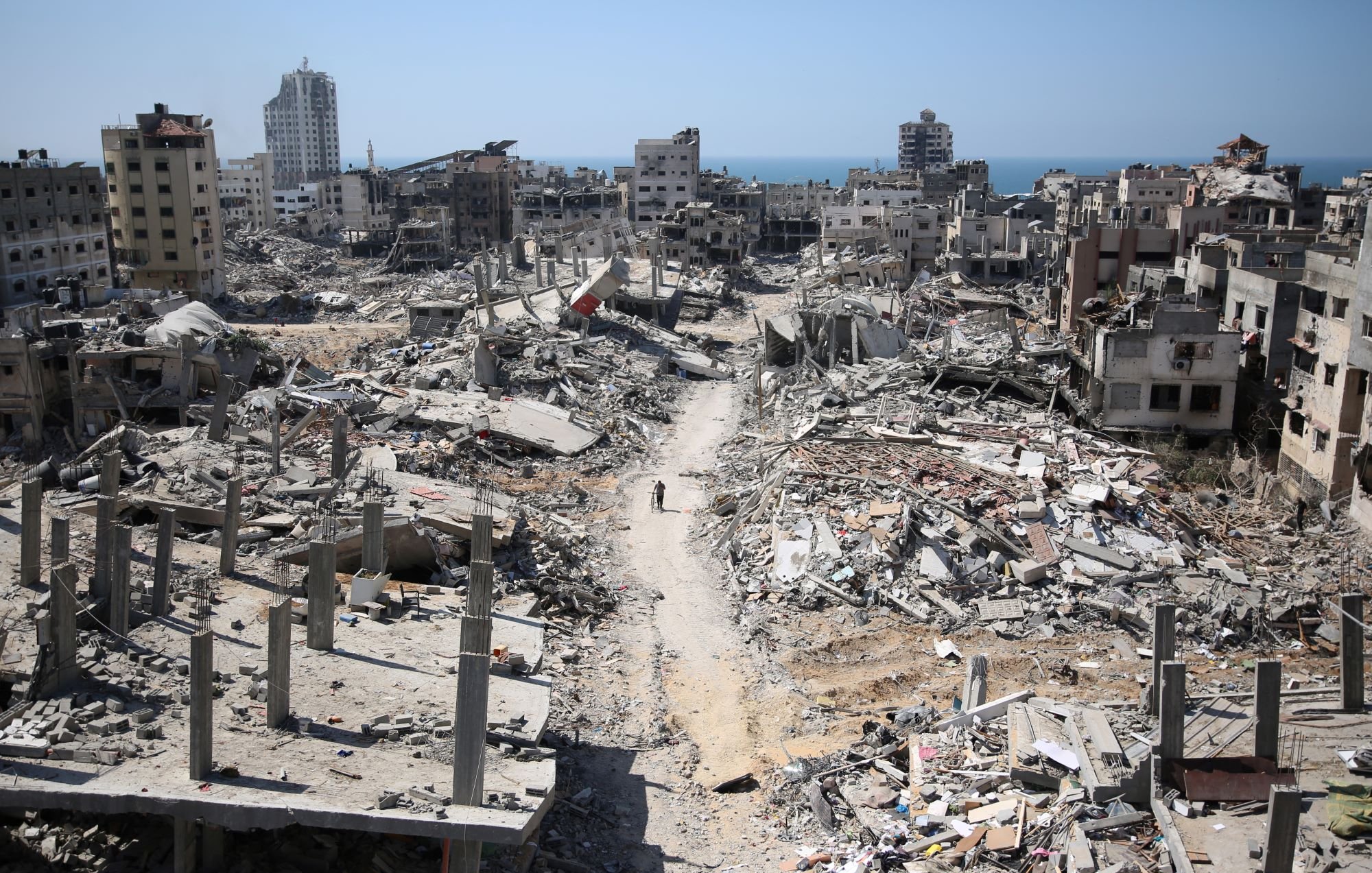 تصویر نشاندهنده یک منطقه ویران شده است که ساختمانهای تخریبشده و خرابهها در آن دیده میشوند. در پسزمینه، دریا نمایان است و آسمان آبی رنگ است. در این صحنه، یک مسیر خاکی به چشم میخورد که از میان ویرانهها عبور میکند و در انتهای آن، فردی در حال حرکت است. این تصویر حس غم و ویرانی را منتقل میکند و نشاندهنده آسیبهای ناشی از جنگ یا بلایای طبیعی است.