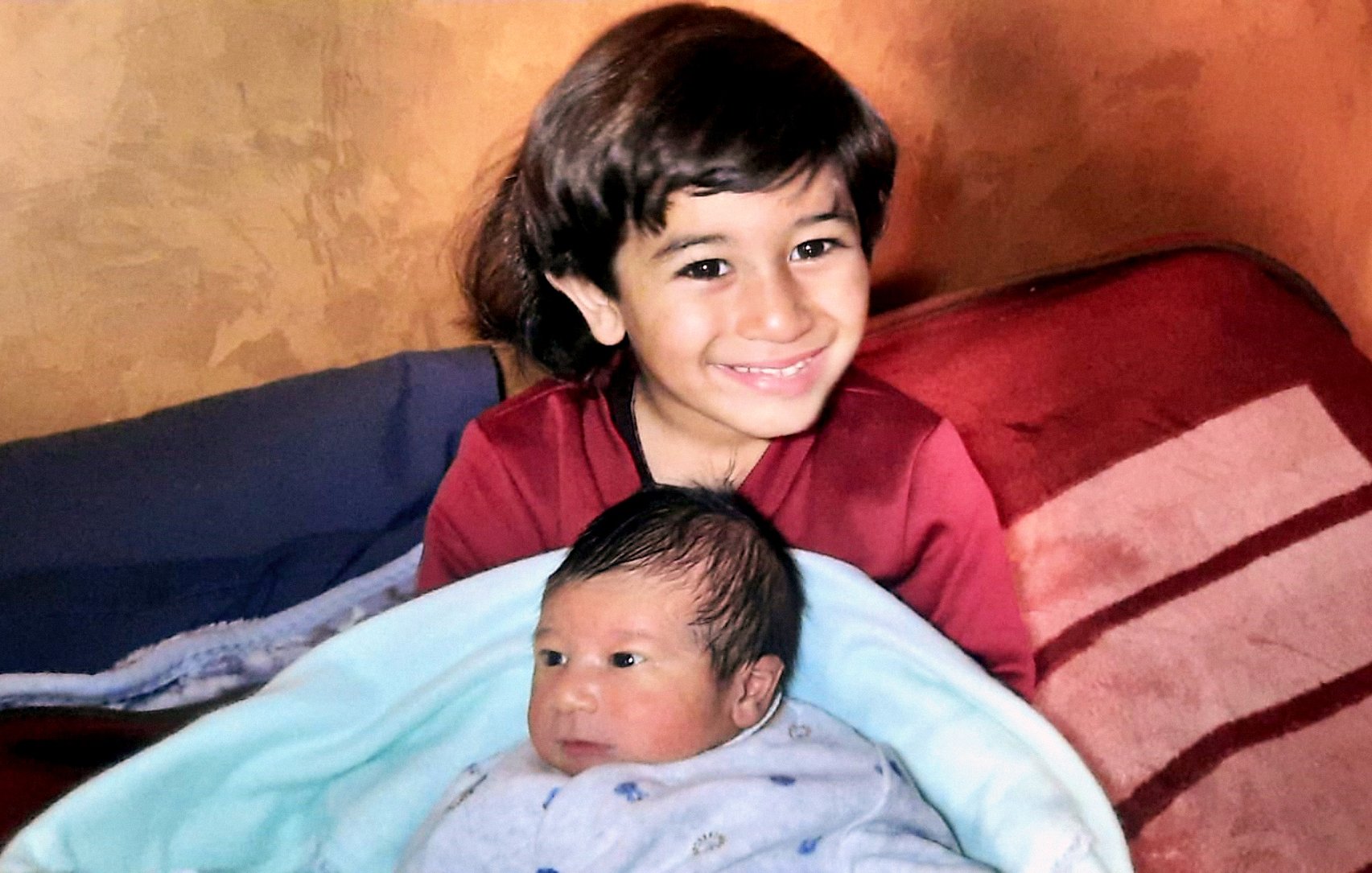 تصویر یک پسر بچهی حدود ۵ ساله را نشان میدهد که لبخند میزند و در آغوش او یک نوزاد خوابیده است. پسر بزرگتر لباس قرمز پوشیده و نوزاد در یک پتوی آبی پیچیده شده است. پسزمینه تصویر رنگی گرم دارد.