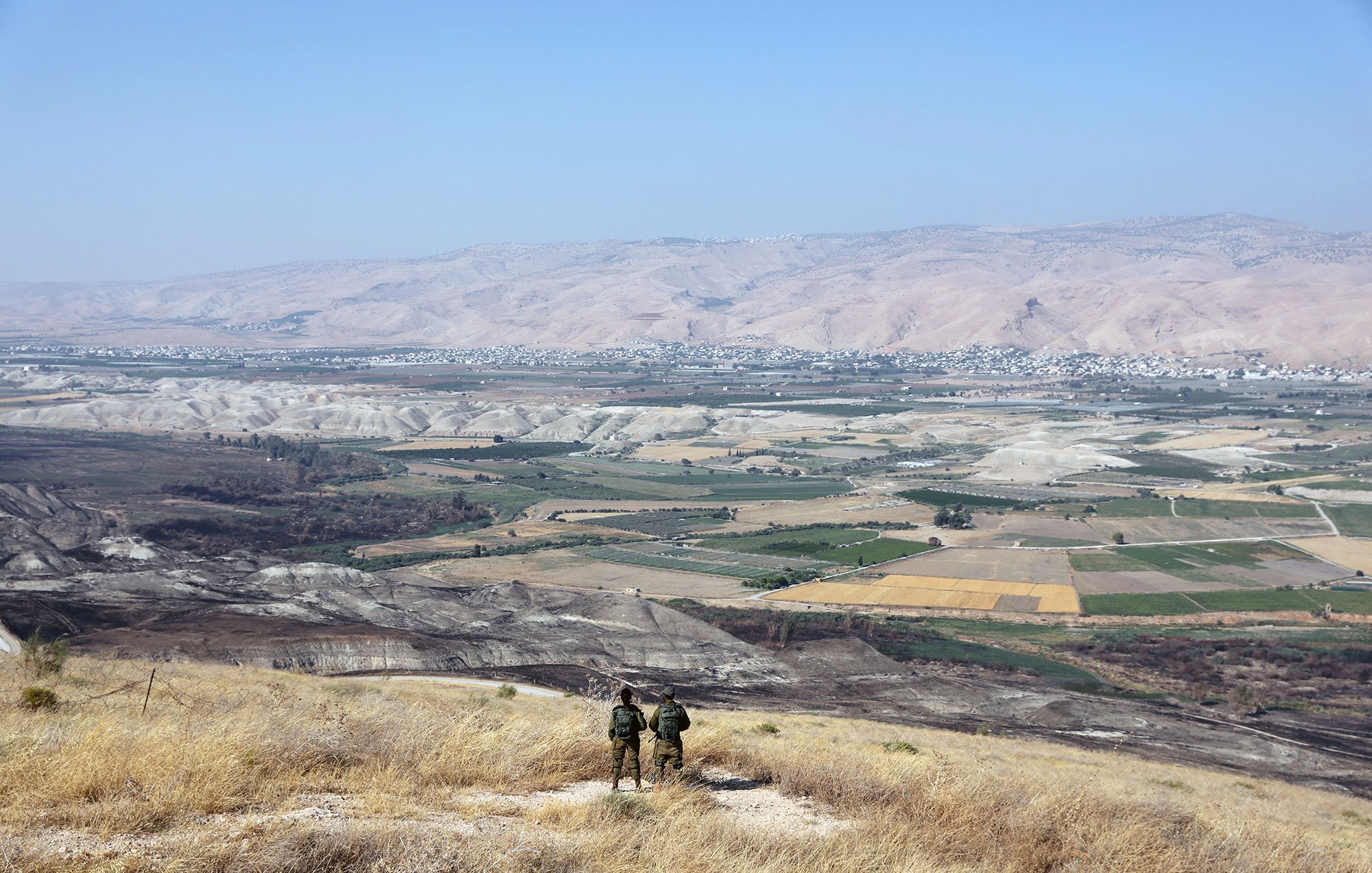 در این تصویر، دو نفر در حال ایستادن بر روی یک تپه یا ارتفاع هستند و به منظرۀ وسیع و زیبای زیر پای خود نگاه میکنند. در زیر، دشتها و زمینهای کشاورزی با رنگهای مختلف دیده میشوند. دامنههای کوهها در دوردست قرار دارند و آسمان آبی و صاف به چشم میخورد. فضا به نظر آرام و دلنشین میرسد و نشاندهندهٔ مناظر طبیعی و انسانی در منطقهای کوهستانی است.