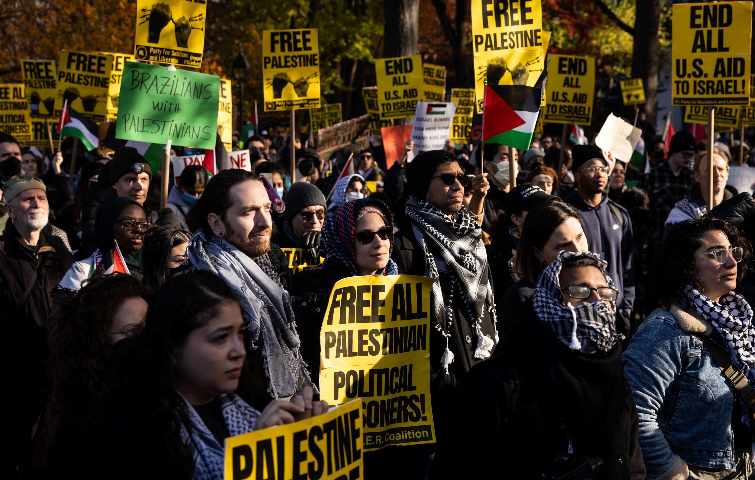 در این تصویر یک تجمع اعتراضی را مشاهده میکنیم که افراد زیادی در آن حضور دارند. شرکتکنندگان با پلاکاردهایی در دست دارند که بر روی آنها نوشتههای مربوط به آزادی فلسطین و حمایت از فلسطینیها دیده میشود. برخی از حاضرین نیز حول گردن خود شالهایی دارند که به نمادهای فرهنگی اشاره دارد. جو کلی تجمع به سمت حمایت از حقوق فلسطینیها و درخواست برای آزادی آنهاست. در پسزمینه، تعدادی درخت و ساختمانها دیده میشوند که نشاندهنده یک مکان عمومی است.