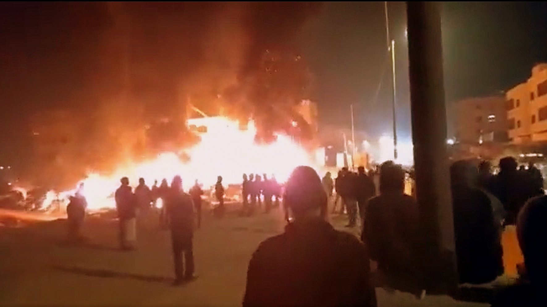 در این تصویر، ظاهراً افرادی در یک صحنه شبانه به سوی یک آتش بزرگ در حال تماشای آن هستند. شعلههای آتش به شدت در حال سوزاندن یک ساختمان یا ساختار دیگر هستند و دود سیاهی به سمت آسمان بلند میشود. مردم در گروههای مختلف در پسزمینه ایستادهاند و به سمت محل آتش نگاه میکنند. احتمالاً این صحنه به یک حادثه یا اعتراض مربوط میشود. روشنایی آتش فضای اطراف را درخشان کرده است.