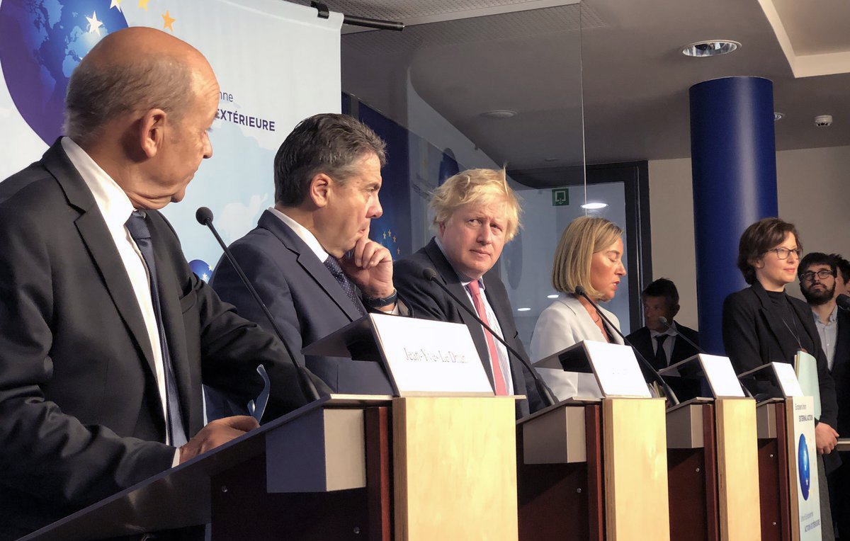 در این تصویر، گروهی از سیاستمداران در حال برگزاری یک کنفرانس خبری یا نشست مطبوعاتی هستند. چندین نفر بر روی چهار پایه ایستادهاند که هر کدام به یک میکروفن مجهز هستند. در میان آنها مردی با موهای بلوند و کوتاه به چشم میخورد که به نظر میرسد یکی از افراد کلیدی این نشست است. سایر افراد نیز در حالتهای مختلف ایستاده و به صحبتهای یکدیگر گوش میدهند. پسزمینه تصویر شامل یک صفحه نمایش یا دیوار با نشانههای مربوط به جلسه است. این تصویر به فضای رسمی و دیپلماتیک اشاره دارد.