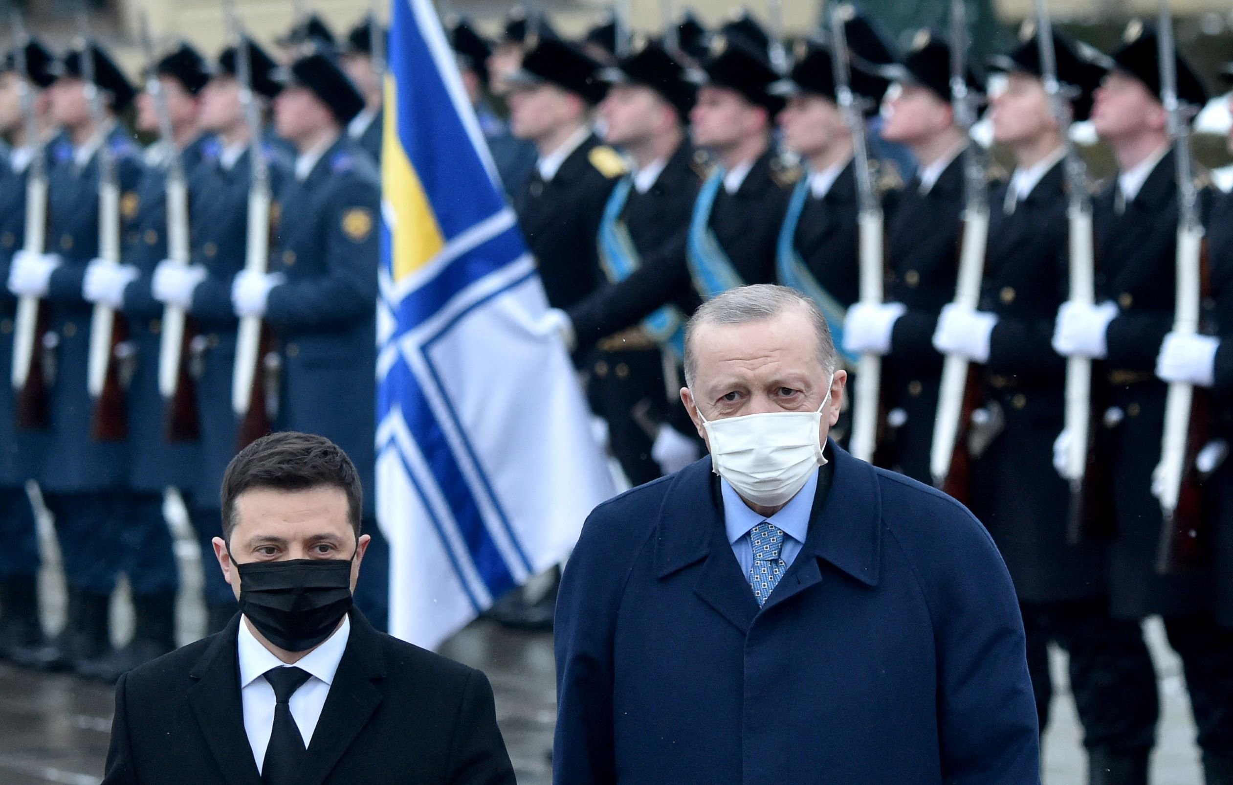 در تصویر، دو مرد در پیشزمینه دیده میشوند که هر دو ماسک زدهاند. یکی از آنها ظاهراً مقام رسمی است و دیگری نیز به نظر میرسد که رهبر یک کشور باشد. در پسزمینه، یک رژه نظامی در حال برگزاری است و سربازان با یونیفورمهای رسمی و پرچمهایی در دست ایستادهاند. فضای کلی تصویر رسمی و جدی به نظر میرسد.