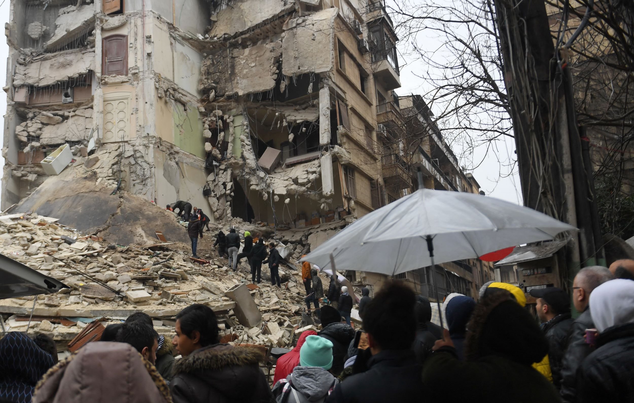 این تصویر یک صحنه ویران شده از یک زمینلرزه را نمایش میدهد. در تصویر، تعدادی از افرادی که به سمت محل خرابی رفتهاند، دیده میشوند. ساختمانها به شدت آسیب دیده و برخی از دیوارها و تکیهگاهها فرو ریختهاند. همچنین چند نفر با چتر در حال نظارت و کمک به دیگران هستند. مردم با نگران و متعجب به این صحنه نگاه میکنند، و احساس غم و ناراحتی در فضا حاکم است.