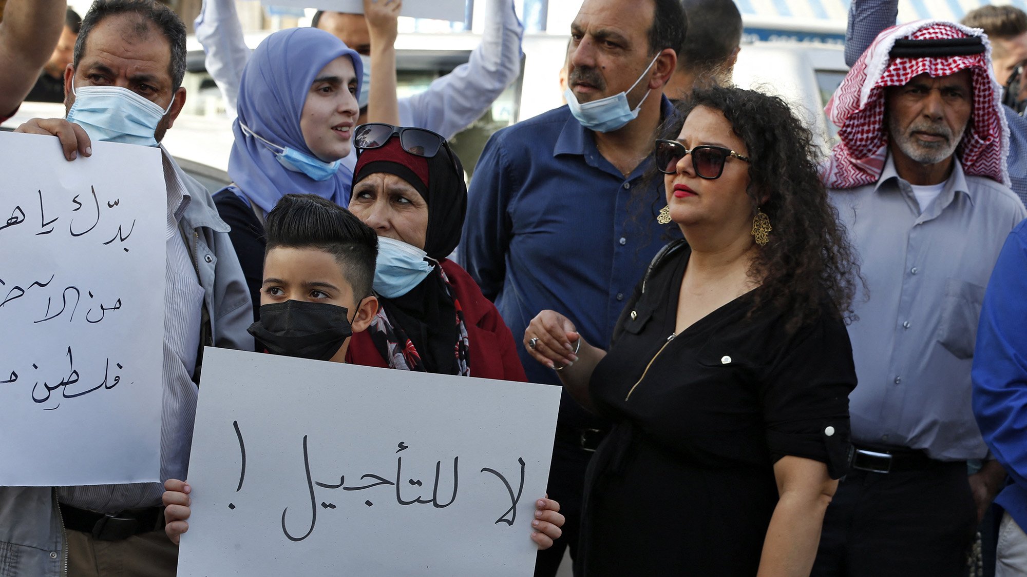در این تصویر، گروهی از افراد در حال تظاهرات هستند. برخی از آنها ماسک به صورت دارند و تابلوهایی در دست دارند. یکی از تابلوها حاوی جملهای است که به زبان عربی نوشته شده و به معنای "نه به تاخیر!" است. افراد در تصاویر شامل زنان و مردانی با پوششهای مختلف هستند، و برخی از آنها با چهرههای جدی و مصمم به بیان خواستههای خود پرداختهاند. فضای کلی نشاندهندهی اعتراض و تلاش برای بیان نظرات و مطالبات است.