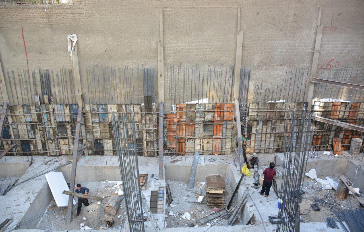 تصویر یک محل construction (ساخت و ساز) را نشان میدهد. در این تصویر، یک سازه بتونی در حال انجام است که دیوارهای آن با میلههای فولادی و قالبهای چوبی محصور شدهاند. عدهای از کارگران در حال فعالیت و جابجایی مصالح هستند. زمین پروژه نیز برخی مصالح و تجهیزات ساختمانی مانند تخته، میلههای فولادی و ابزارهای دیگر را نشان میدهد. دیوار پشتی با یک سطح صاف و عاری از هرگونه تزئینات، نمایان است.