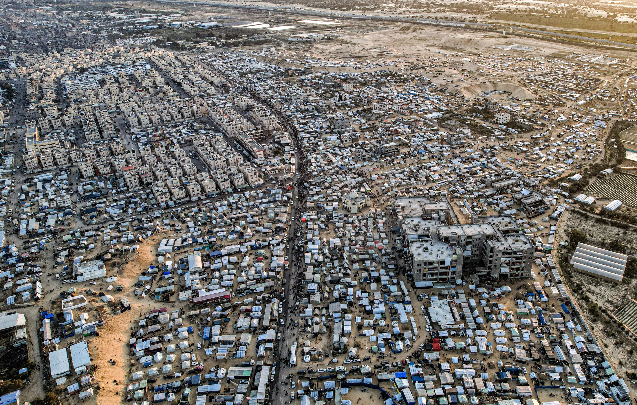 این تصویر نمای هوایی از یک منطقه شلوغ و پرجمعیت است. در اینجا تعداد زیادی ساختمان و خانه های کوچک دیده میشود که در کنار هم قرار گرفتهاند. خیابانهایی که به نظر میرسد مردم در آن تردد دارند، میانه تصویر را تقسیم کردهاند. همچنین، در برخی نواحی، چادرها و سازههای موقت دیده میشوند که نشاندهنده زندگی در شرایط سخت و غیرفرهنگین است. این تصویر، وضعیتی را به نمایش میگذارد که بیانگر چالشهای اجتماعی و اقتصادی است.