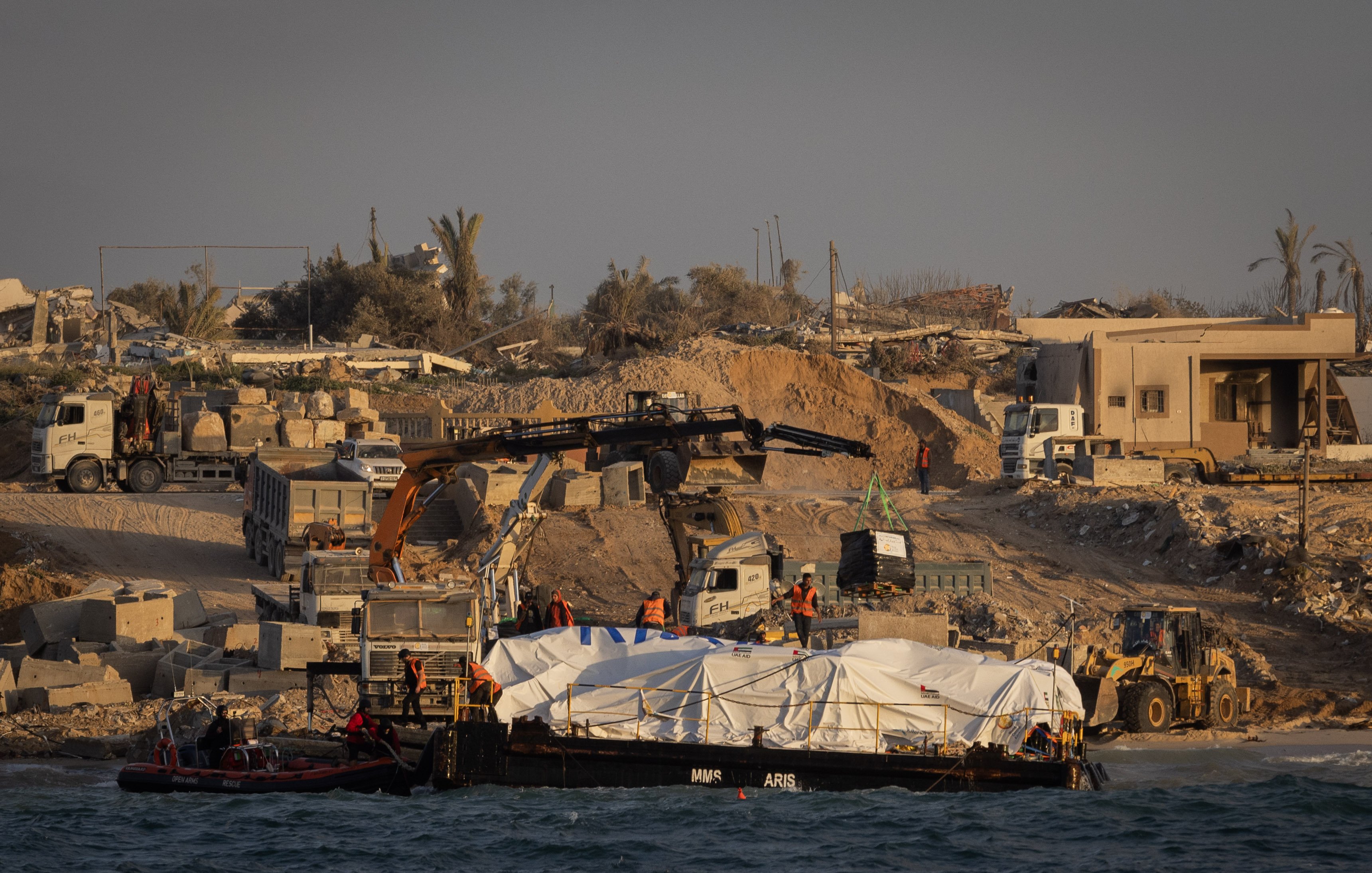 در این تصویر، یک صحنه ساختمانی در کنار دریا دیده میشود. در ساحل، تعدادی ماشین سنگین و جرثقیل در حال کار هستند. یک قایق روی آب قرار دارد که زیر آن یک پوشش سفید رنگ قرار دارد. پسزمینه شامل ساختمانهای نیمهکار و تخریب شده و چند درخت نخل است. نور خورشید به صورت گرم بر روی این صحنه تابیده و حال و هوای آن را به تصویر کشیده است.