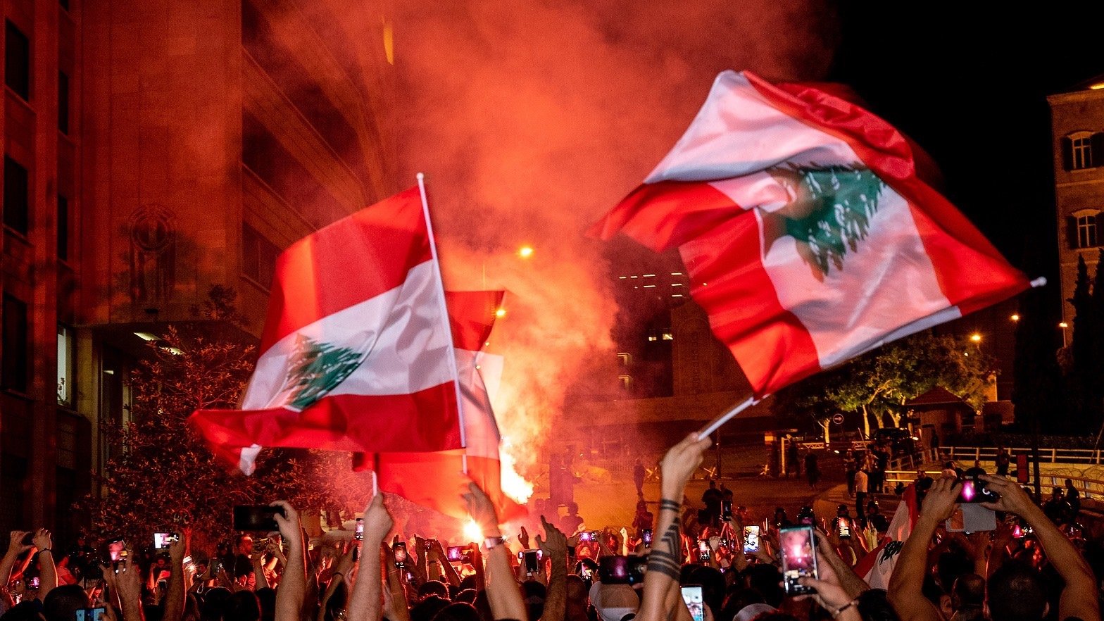 در تصویر، جمعیتی از مردم در یک تظاهرات شبانه دیده میشوند. افراد با پرچمهای لبنان در دست، در حال شادی و ابراز احساسات هستند. یک شعله آتش بزرگ در پسزمینه به روشنایی فضای شب افزوده و به جو فعالیت و هیجان دامن میزند. تعداد زیادی از افراد گوشیهای هوشمند خود را برای ثبت لحظات بالا بردهاند. این صحنه نمایانگر شور و شوق اجتماعی و همبستگی مردم است.