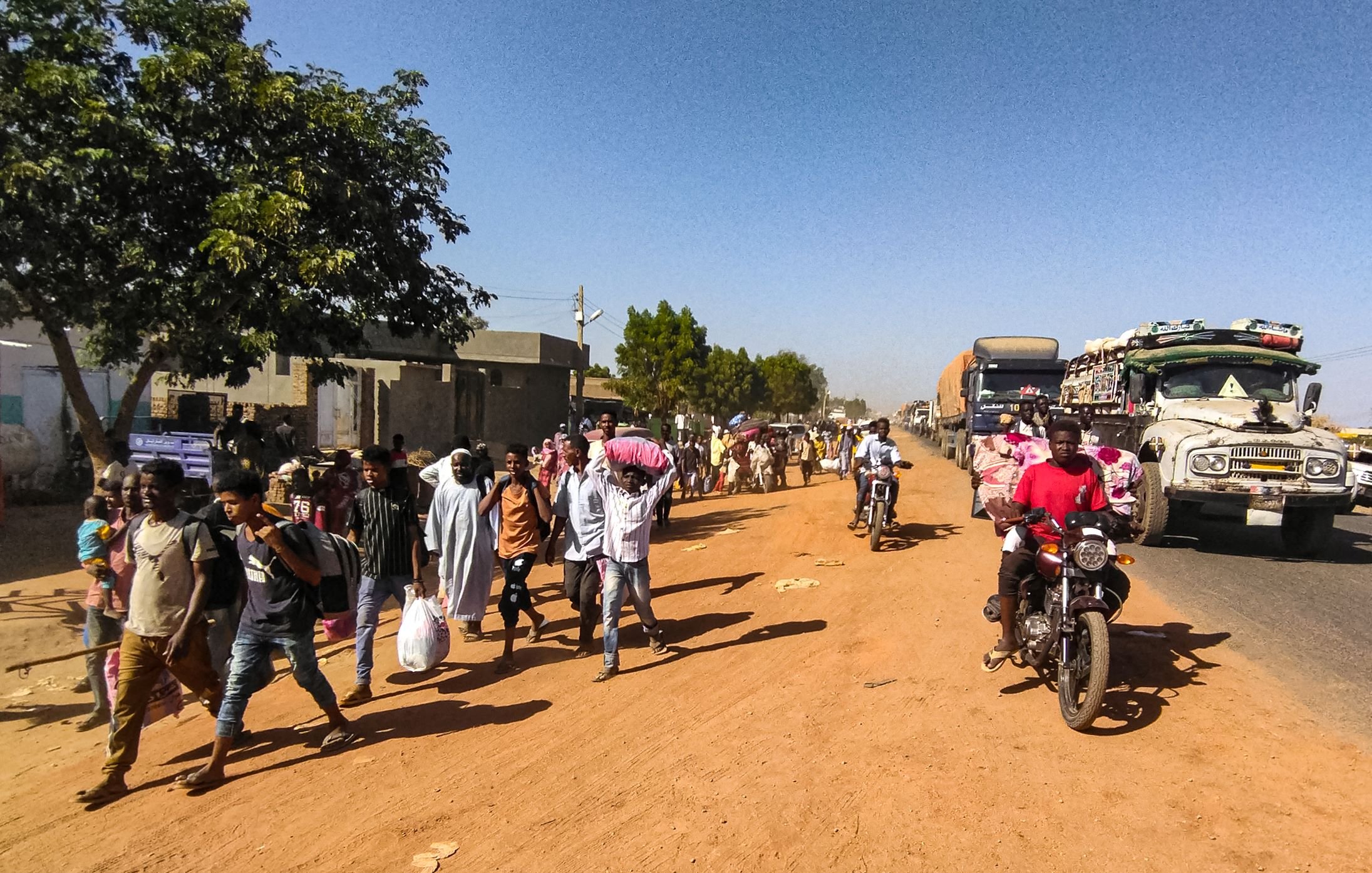 در این تصویر، یک خیابان شنی و پرجنبوجوش دیده میشود. در سمت چپ، تعدادی درخت و ساختمانهای ساده قرار دارند. گروهی از مردم در حال پیادهروی هستند و برخی از آنها کیسههایی به دوش دارند. در سمت راست، چندین خودرو و موتورسیکلت در حال تردد هستند. فضای عمومی پر از فعالیت و زندگی به نظر میرسد.