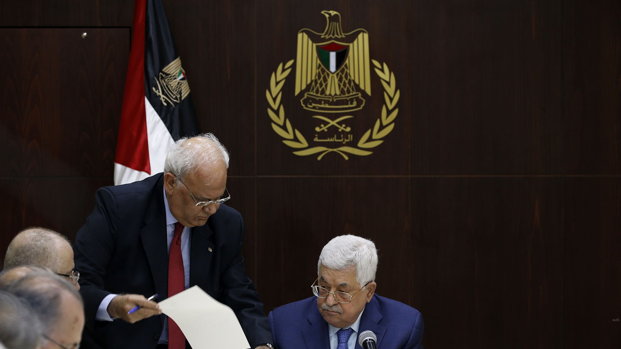 در این تصویر، یک جلسه رسمی در حال برگزاری است. در مرکز تصویر، مردی به نام محمود عباس نشسته است که به نظر میرسد در حال مطالعه مدارک یا مستندات است. افراد دیگری نیز در اطراف او نشستهاند. یکی از آنها به سمت عباس خم شده و به او مدرکی نشان میدهد. در پسزمینه، پرچم فلسطین و نماد رسمی مرتبط با دولت فلسطین دیده میشود، که به فضایی رسمی و سیاسی اهمیت میدهد. میز گردی که آنها دور آن نشستهاند، با لوازم اداری و بطریهای آب تزیین شده است.