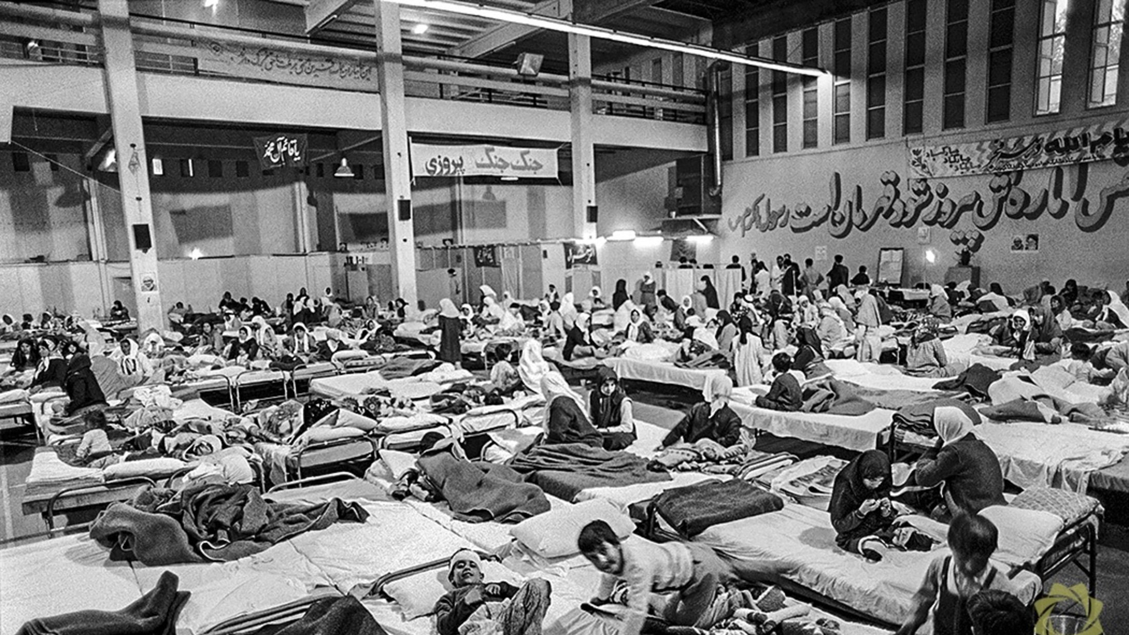 این تصویر یک فضای داخلی را نشان میدهد که پر از تختهای بیمارستانی است. افراد مختلفی در حالتهای مختلف دراز کشیدهاند، برخی در حال استراحت و برخی دیگر در حال حرکت یا صحبت با یکدیگر هستند. محیط به نظر شلوغ و پر از فعالیت میرسد، و پوشش بیشتر افراد ساده و شامل چادر یا پتو است. دیوارها به نوعی دکور شدهاند، و فضای کلی شاید به یک مرکز درمانی یا پناهگاه اشاره دارد.