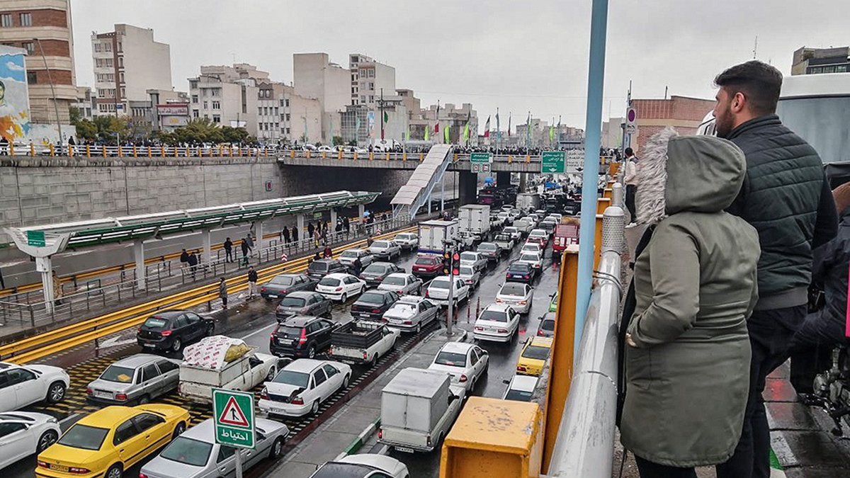تصویر نشاندهنده یک صحنه شلوغ از ترافیک در یک خیابان شهری است. در سمت چپ تصویر، افرادی در حال تماشا از بالا ایستادهاند. در پایین، خطوط خودروها به وضوح قابل مشاهده است که نشاندهنده تجمع و ترافیک سنگین است. بقیه عناصر شهری مانند ساختمانها و علائم راهنمایی نیز در پسزمینه دیده میشوند. هوای ابری و احتمالا بارانی به دید فضا و حس و حال تصویر اضافه میکند.
