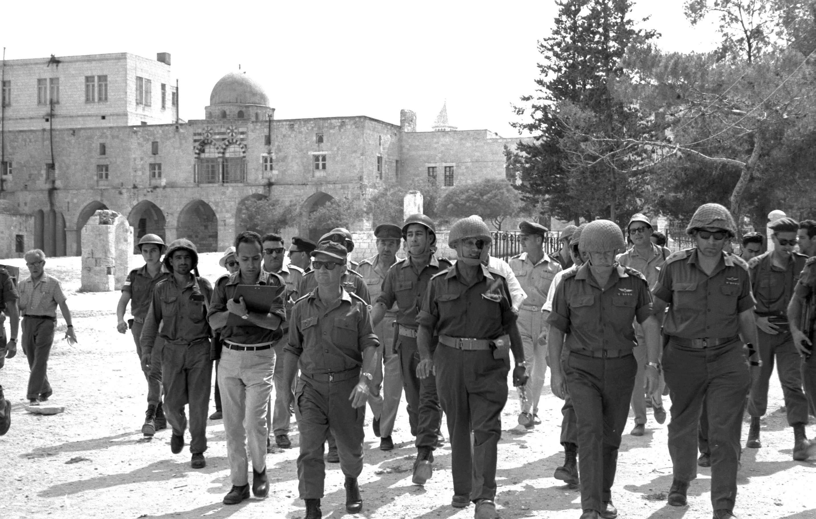 در این تصویر، گروهی از نظامیان و مأموران امنیتی در حال راه رفتن به سمت دوربین هستند. آنها لباسهای نظامی و کلاههای مخصوص به سر دارند. پسزمینه تصویر، شامل ساختمانهای سنگی قدیمی و برخی درختان است. به نظر میرسد این تصویر در یک منطقه تاریخی یا باستانی گرفته شده باشد. نور خورشید به وضوح بر روی صحنه میتابد و فضایی گرم به تصویر میبخشد.