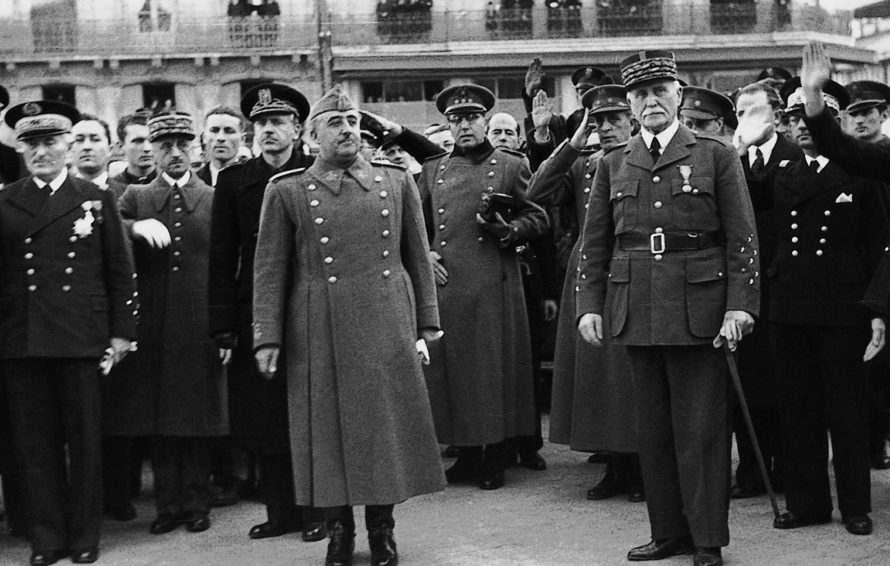 این تصویر سیاه و سفید یک جمعیت نظامی را نشان میدهد که شامل تعدادی افسر و مقامهای عالیرتبه است. در مرکز تصویر، دو نفر ایستادهاند که به نظر میرسد شخصیتهای مهمی هستند. یکی از آنها در یک کت بلند و با نشانهای نظامی دیده میشود و دیگری نیز لباسی مشابه به تن دارد. افراد دیگر در پسزمینه به حالت احترام دست خود را بالا بردهاند و به نظر میرسد در حال ادای احترام هستند. فضای کلی تصویر جدی و رسمی به نظر میرسد.