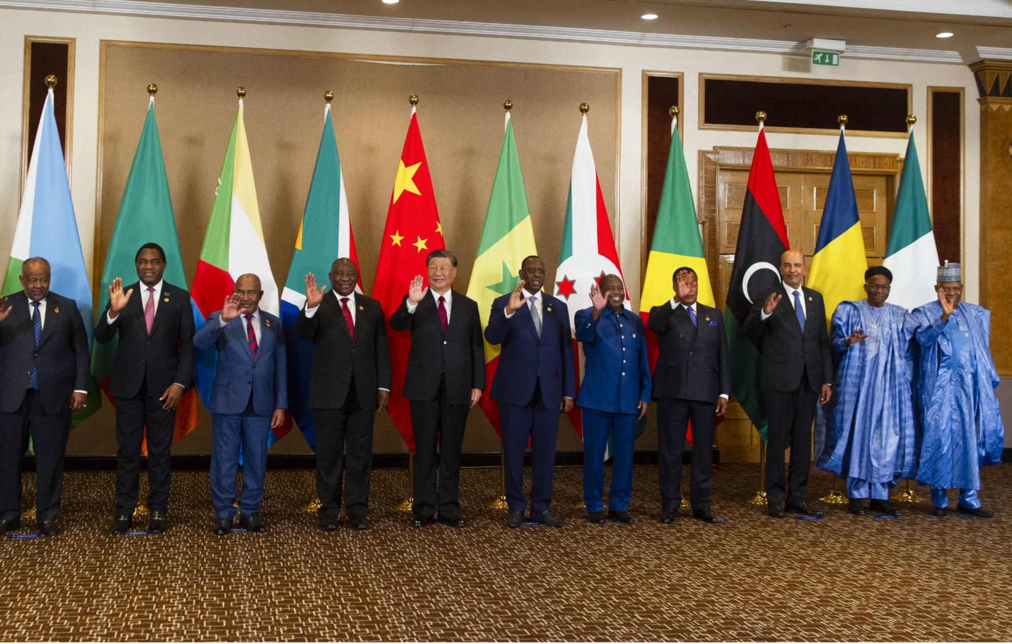 تصویر یک گروه از افراد را نشان میدهد که در کنار هم ایستادهاند و پرچمهای مختلف کشورهایشان در پسزمینه قرار دارد. به نظر میرسد این افراد مقامهای بلندپایه یا رهبران سیاسی هستند و همگی در حال خوشآمدگویی یا دست wave کردن به دوربین هستند. پرچمهای مختلف شامل پرچمهای کشورهای آفریقایی و همچنین پرچمهای چین و سایر کشورها وجود دارد. فضای تصویر رسمی به نظر میرسد و افراد لباسهای رسمی بر تن دارند.
