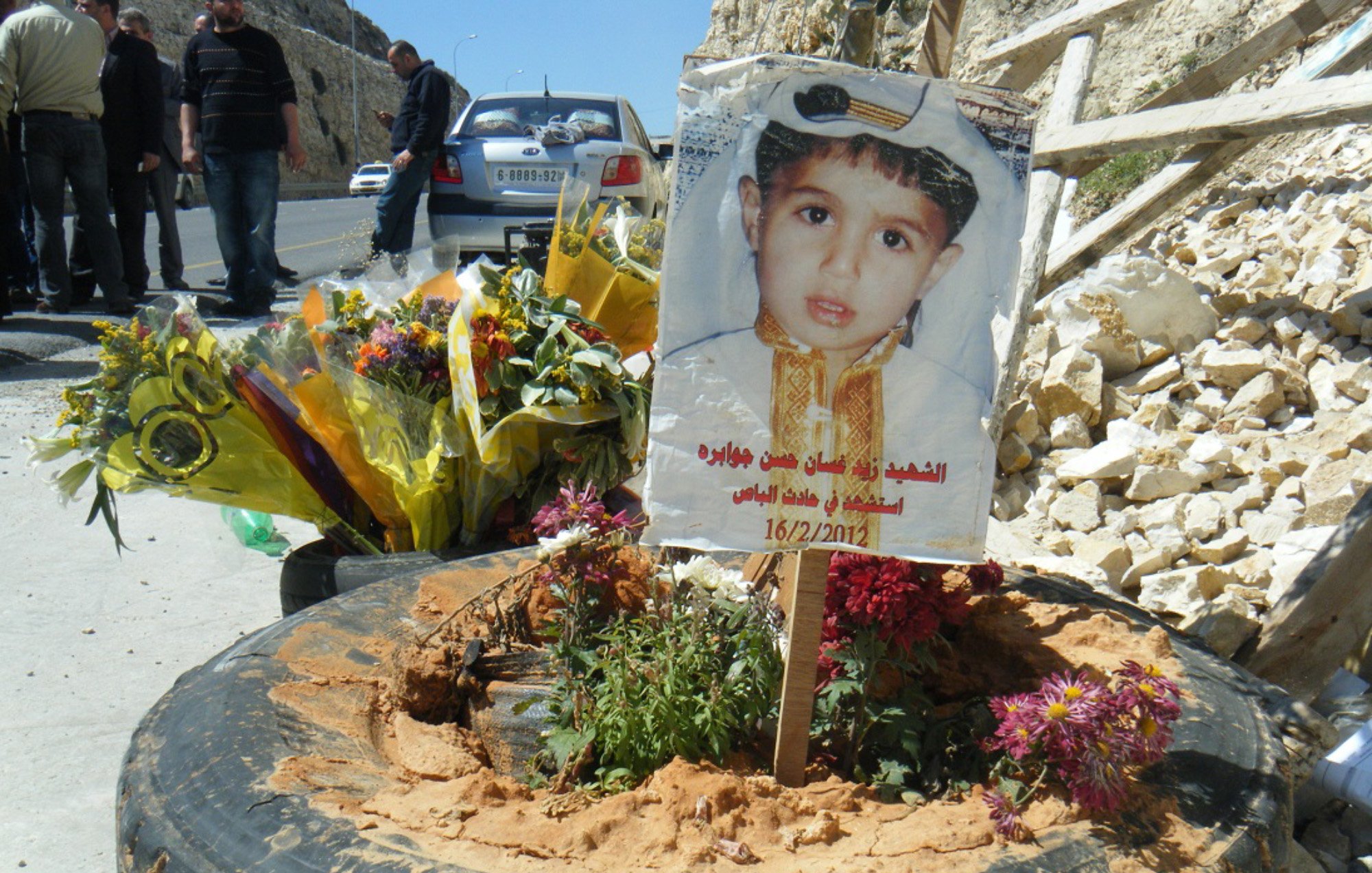 تصویر نشاندهنده یک یادبود است که به یاد یک کودک ساخته شده است. در مرکز تصویر، یک دسته گل بزرگ و زیبا قرار دارد و در کنار آن، تابلویی با عکس کودک وجود دارد که لباس خاصی به تن دارد و به تاریخ 16/2/2012 اشاره شده است. زمین اطراف یادبود با شن و گلهای رنگارنگ تزئین شده و در پسزمینه، تعدادی از افراد به نظر میرسد که در حال حضور در آن مکان هستند. تصویر احساسات عمیق و یادآوری را منتقل میکند.