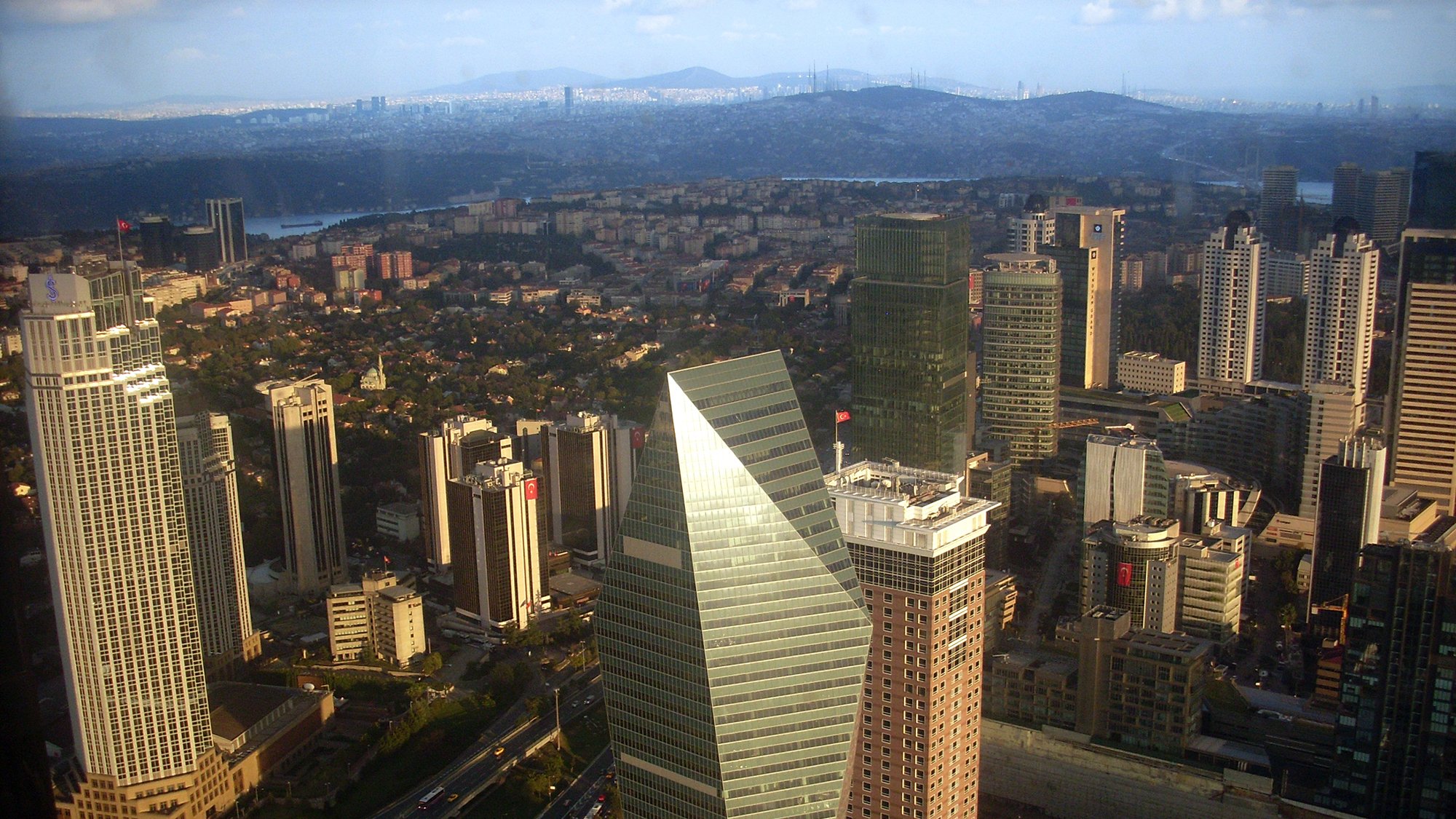 این تصویر نمایی از یک شهر مدرن است که در آن ساختمانهای بلند و شیک مشاهده میشوند. در مرکز تصویر یک ساختمان با طراحی متمایز و زاویهدار وجود دارد که بهنظر میرسد نماد یکی از بانکها باشد. دیگر ساختمانها نیز به ارتفاع و تنوع طراحی در اطراف آن قرار دارند. فضای سبز و خیابانهای در حال تردد نیز در بخشهای پایین تصویر قابل مشاهده است. آسمان آبی با ابرهای خفیف به زیبایی تصویر افزوده است.