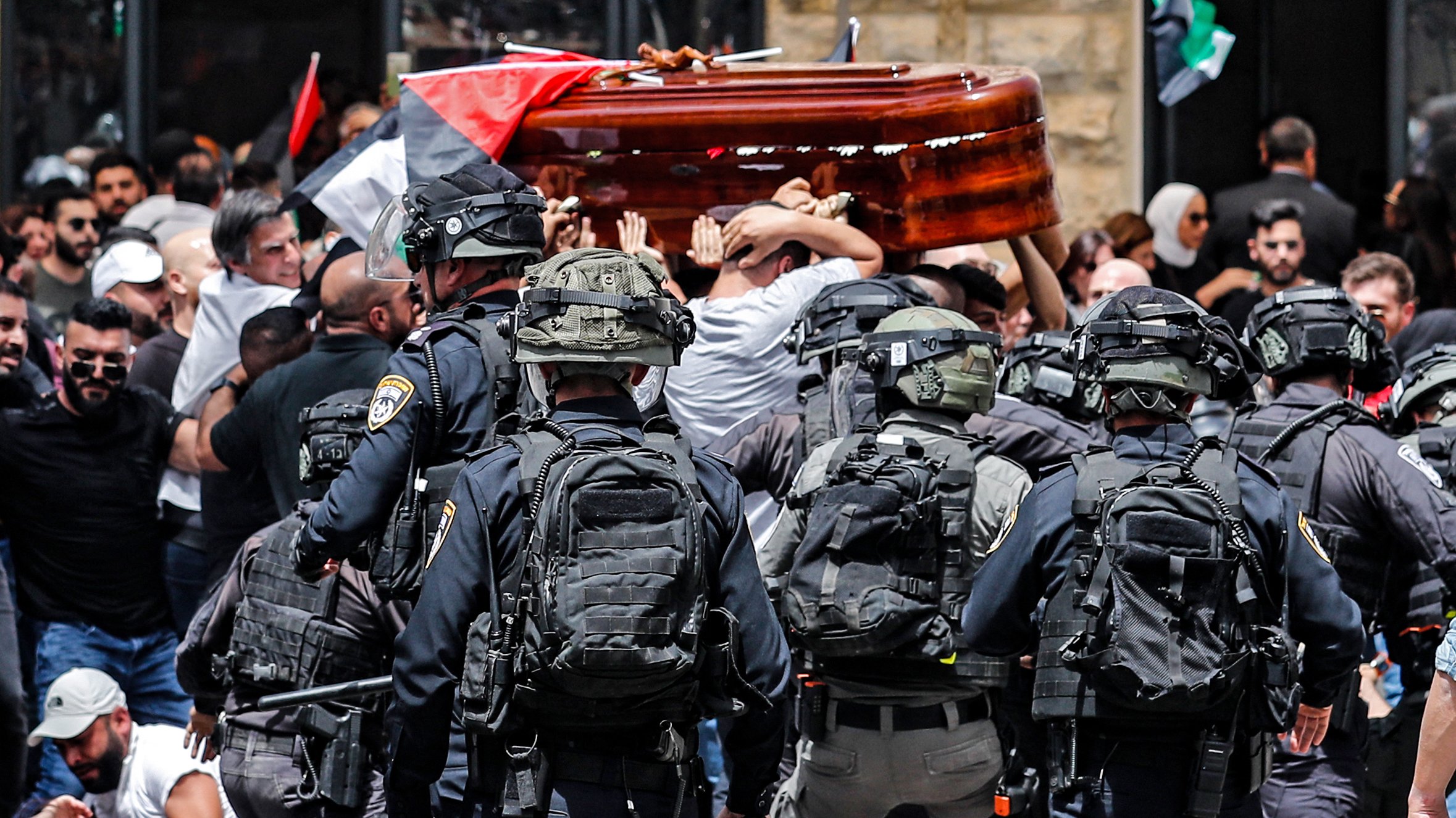 در این تصویر، گروهی از مردم در حال حمل یک تابوت هستند و در اطراف آنها تعدادی نیروی پلیس با تجهیزات نظامی دیده میشود. مردم در حال تجمع و اعتراض هستند و پرچمهایی را در دست دارند. به نظر میرسد این صحنه در یک فضای پرازدحام و تنشزای اجتماعی اتفاق میافتد. واکنشهای متفاوتی در چهرهها و حرکات افراد مشهود است.