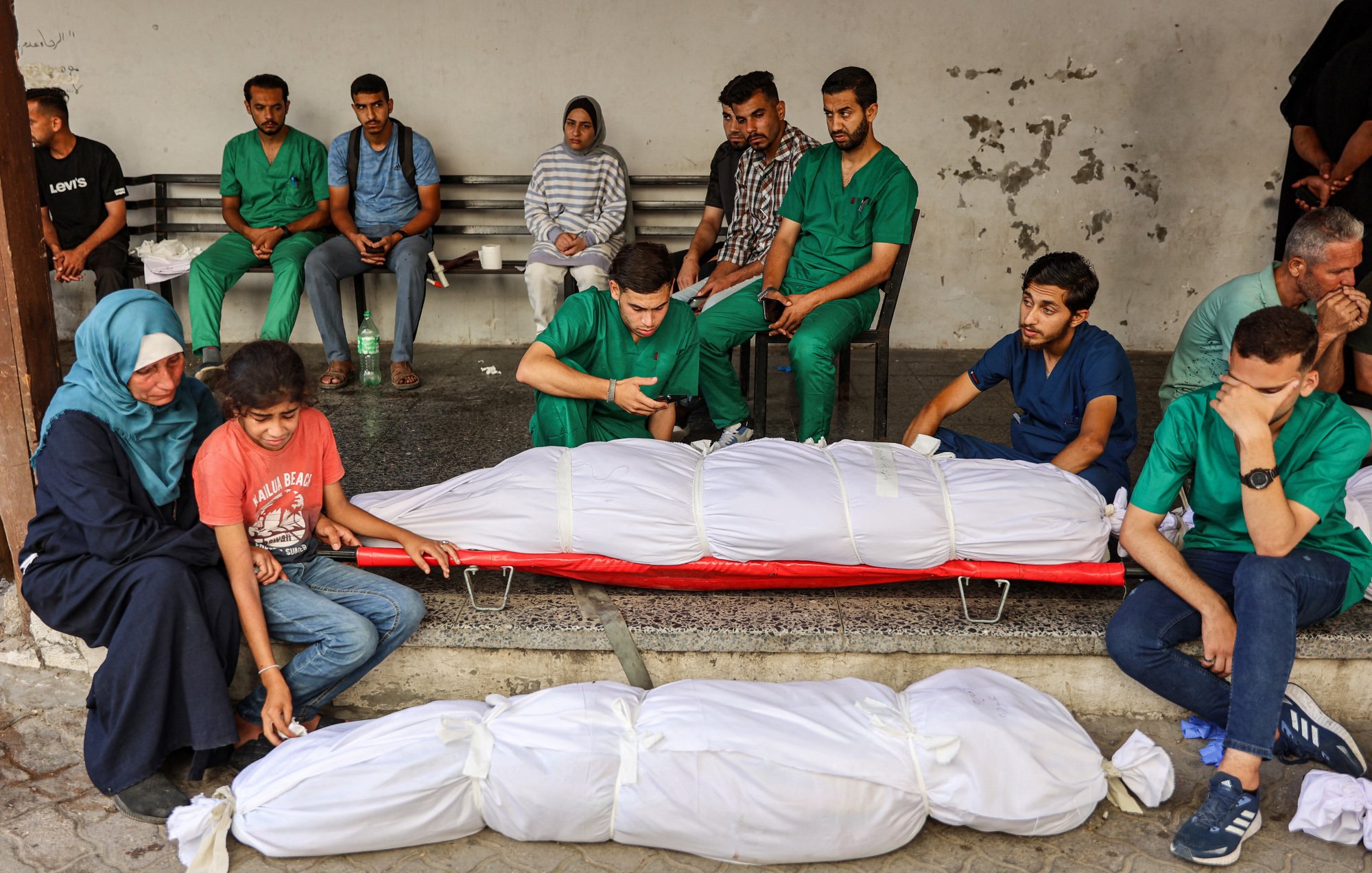 در این تصویر، تعدادی از افراد در یک مکان عمومی نشستهاند. در جلو، دو بدن پوشیده شده در پارچههای سفیدی قرار دارند. افراد حاضر در تصویر با ظاهری نگران و غمگین به نظر میرسند. برخی از آنها لباسهای پزشکی به تن دارند و به وضوح تحت تأثیر شرایط عاطفی و وضعیت حاضر قرار گرفتهاند. فضا به طور کلی حاکی از یک وضعیت بحرانی و دردناک است.