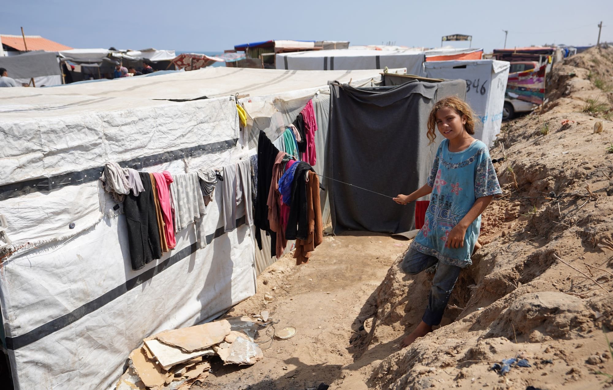 تصویر یک دختر بچه را در یک محیط غیررسمی نشان میدهد که احتمالاً یک اردوگاه پناهندگان یا محلی با شرایط زندگی سخت است. این دختر در کنار یک راه باریک ایستاده و لباسی ساده به تن دارد. در اطراف او، چادرها و سازههای موقت دیده میشود که لباسهایی به طناب آویزان شدهاند. زمین خاکی و نامنظم به نظر میرسد و فضا یک حس ناامنی را منتقل میکند. هوای آبی در پسزمینه نشان میدهد که این مکان نزدیک به دریا یا یک منبع آب است.