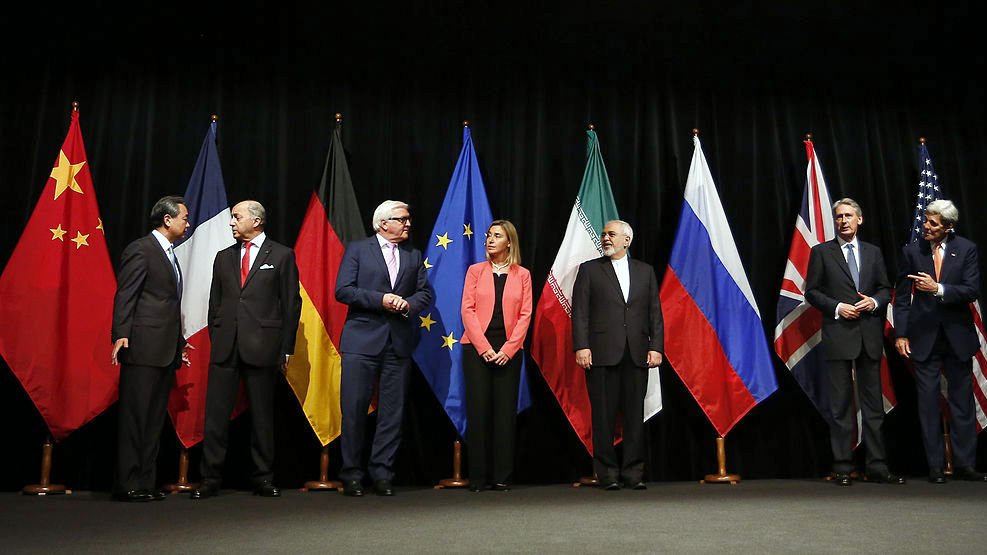 در این تصویر، گروهی از نمایندگان کشورهای مختلف در یک جلسه رسمی ایستادهاند. پرچمهای چین، آلمان، اتحادیه اروپا، ایران، روسیه، بریتانیا و ایالات متحده در پسزمینه حضور دارند. هر یک از افراد در لباسهای رسمی و با حالتهای متفاوتی در صف ایستادهاند، که نشاندهنده اهمیت و جدیت جلسه است. به نظر میرسد آنها در حال گفتوگو و تعامل با یکدیگر هستند.