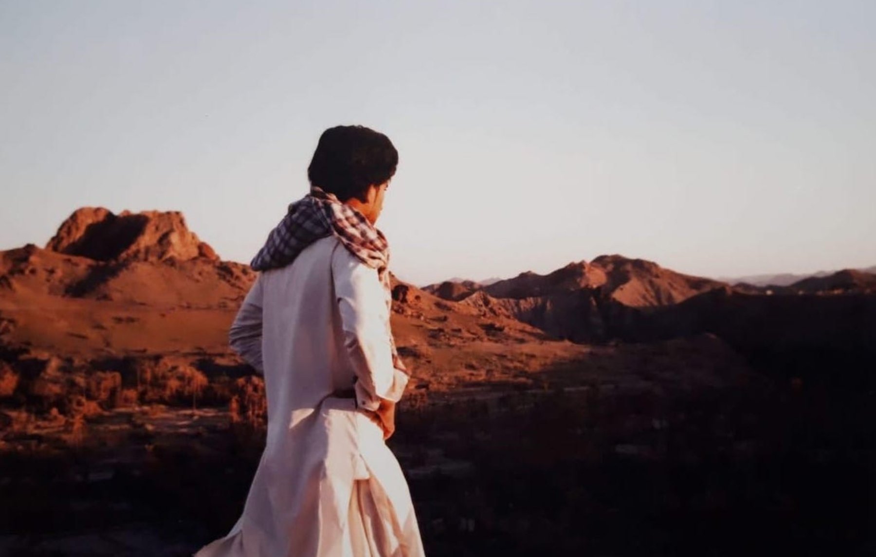در این تصویر، فردی با لباس سنتی و شال بر دوش، به سمت یک چشمانداز طبیعی نگاه میکند. زمینه تصویر شامل کوههای بلند و دشتهای وسیع است که نور ملایم غروب خورشید بر آنها تابیده شده است. رنگهای گرم غروب و سایههای بلند کوهها زیبایی خاصی به صحنه دادهاند. احساس آرامش و سکوت در فضای تصویر به وضوح حس میشود.