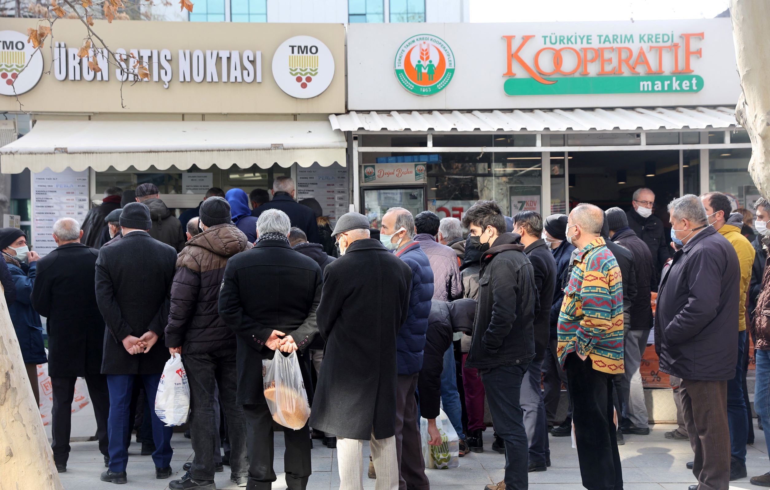 در تصویر، گروهی از مردم، عمدتاً مردان، در حال انتظار در صفی طولانی جلوی یک فروشگاه به نام "Kooperatif market" هستند. بعضی از افراد کیف و بستههای خرید در دست دارند و بیشتر آنها لباسهای گرم پوشیدهاند. در پسزمینه، چند ساختمان و درختان دیده میشود. افراد به نظر میرسد که منتظر خرید محصولاتی از فروشگاه هستند.