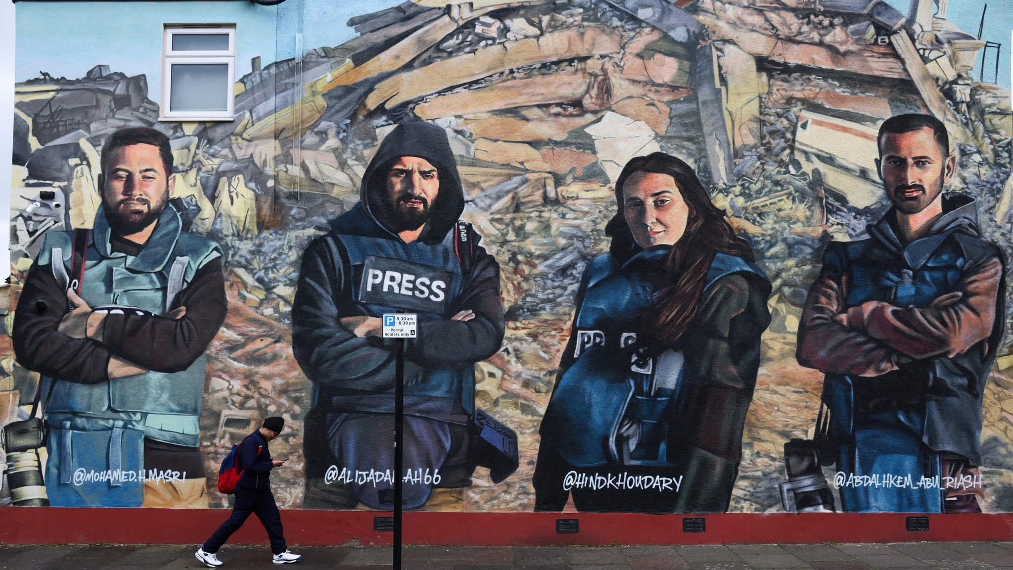 این تصویر یک دیوارنگاری بزرگ را نشان میدهد که چهار نفر را به تصویر کشیده است. این افراد در لباسهای مخصوص خبرنگاری قرار دارند و با ظاهری جدی و ثابت ایستادهاند. در پسزمینه، ویرانههایی دیده میشود که نشاندهندهی یک حادثه یا بحران است. یکی از افرادی که در تصویر قرار دارد، لباس مخصوص "پرس" (خبرنگار) به تن دارد و همگی به دوربین نگاه میکنند. در جلوی دیوارنگاری، فردی در حال عبور است. این تصویر به نظر میرسد که به اهمیت کار خبرنگاران در پوشش اخبار بحرانها اشاره دارد.