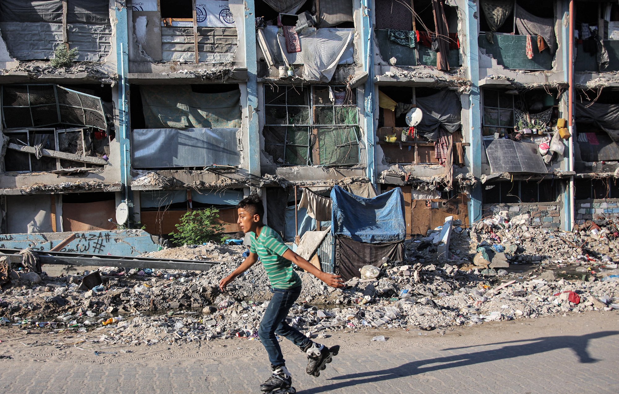 در تصویر یک پسر بچه دیده میشود که با اسکیت روی سطحی صاف در حال حرکت است. در پسزمینه، ساختمانهایی با نمای خراب و آسیبدیده قرار دارند که پنجرههایشان با پارچه و مواد دیگر پوشانده شده است. اطراف ساختمان مملو از زباله و خرابه است که نشاندهنده وضعیت دشوار منطقه است. فضای کلی تصویر حس نابهسامانی و فقر را منتقل میکند.