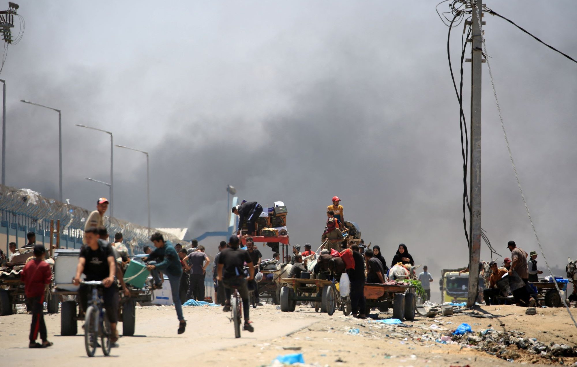 تصویر یک صحنه شهری را نشان میدهد که در آن تعدادی از افراد در حال حرکت هستند. در زمینه، دود سیاهی به آسمان بلند شده که نشاندهنده وجود یک حادثه یا آتشسوزی است. افرادی که در تصویر دیده میشوند، شامل مردان، زنان و کودکانی هستند که برخی از آنها در حال دوچرخهسواری و برخی دیگر در حال حمل بار در کالسکههای دستی هستند. فضای کلی تصویر به نظر شلوغ و پر از فعالیت است.