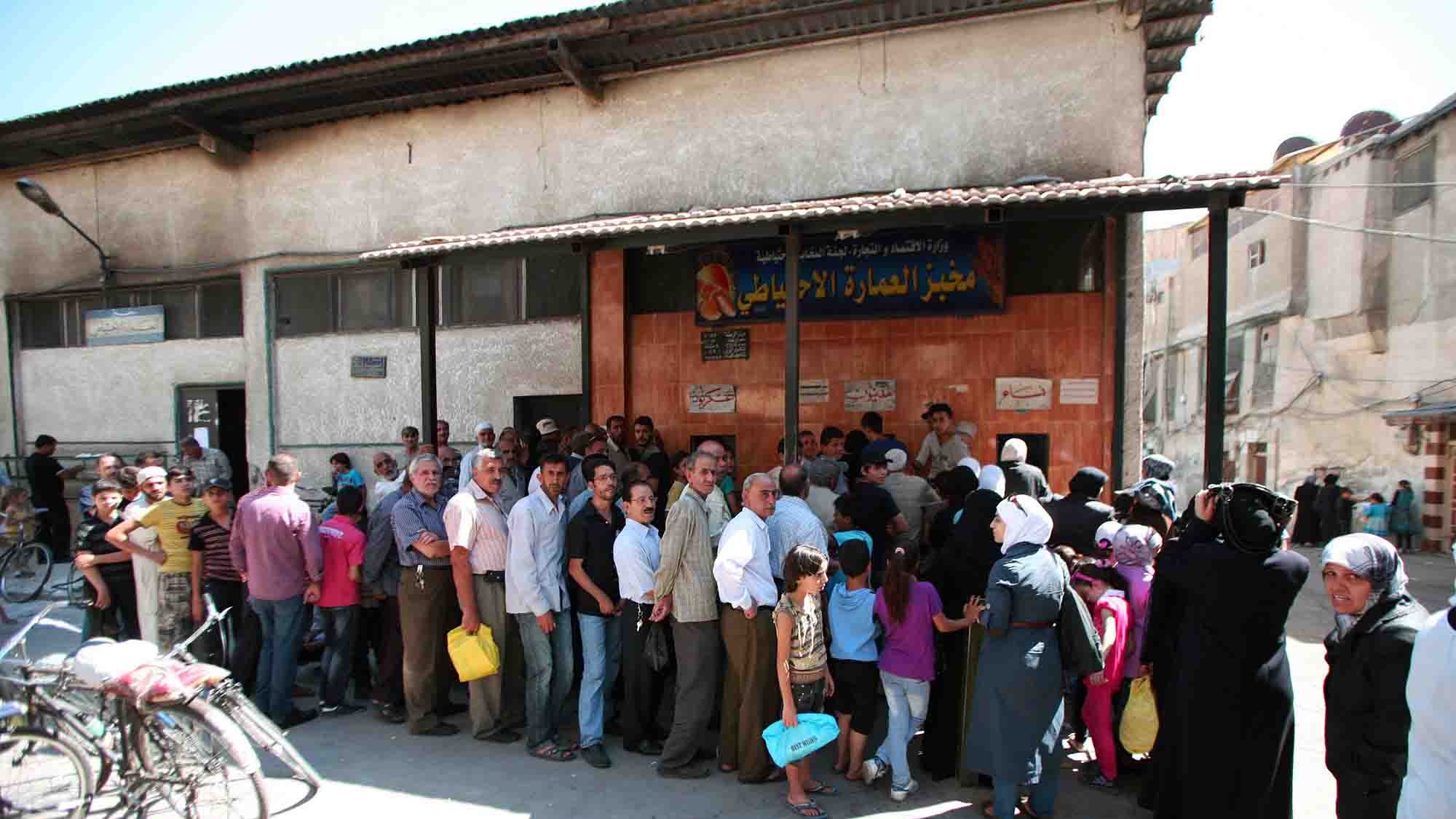 در تصویر، گروهی از مردم به صف ایستادهاند. ساختمان در پسزمینه دارای نمای سادهای است و تابلو بزرگی بالای درب ورودی نصب شده است. افراد در صف شامل مردان و زنانی هستند که برخی از آنها سبدها یا کیسههای خرید را در دست دارند. نور خورشید قوی به تصویر تابیده و فضا شلوغ به نظر میرسد. برخی از افراد به طرف دوربین نگاه میکنند و برخی دیگر مشغول صحبت هستند. دوچرخهها نیز در کنار خیابان به چشم میخورند.