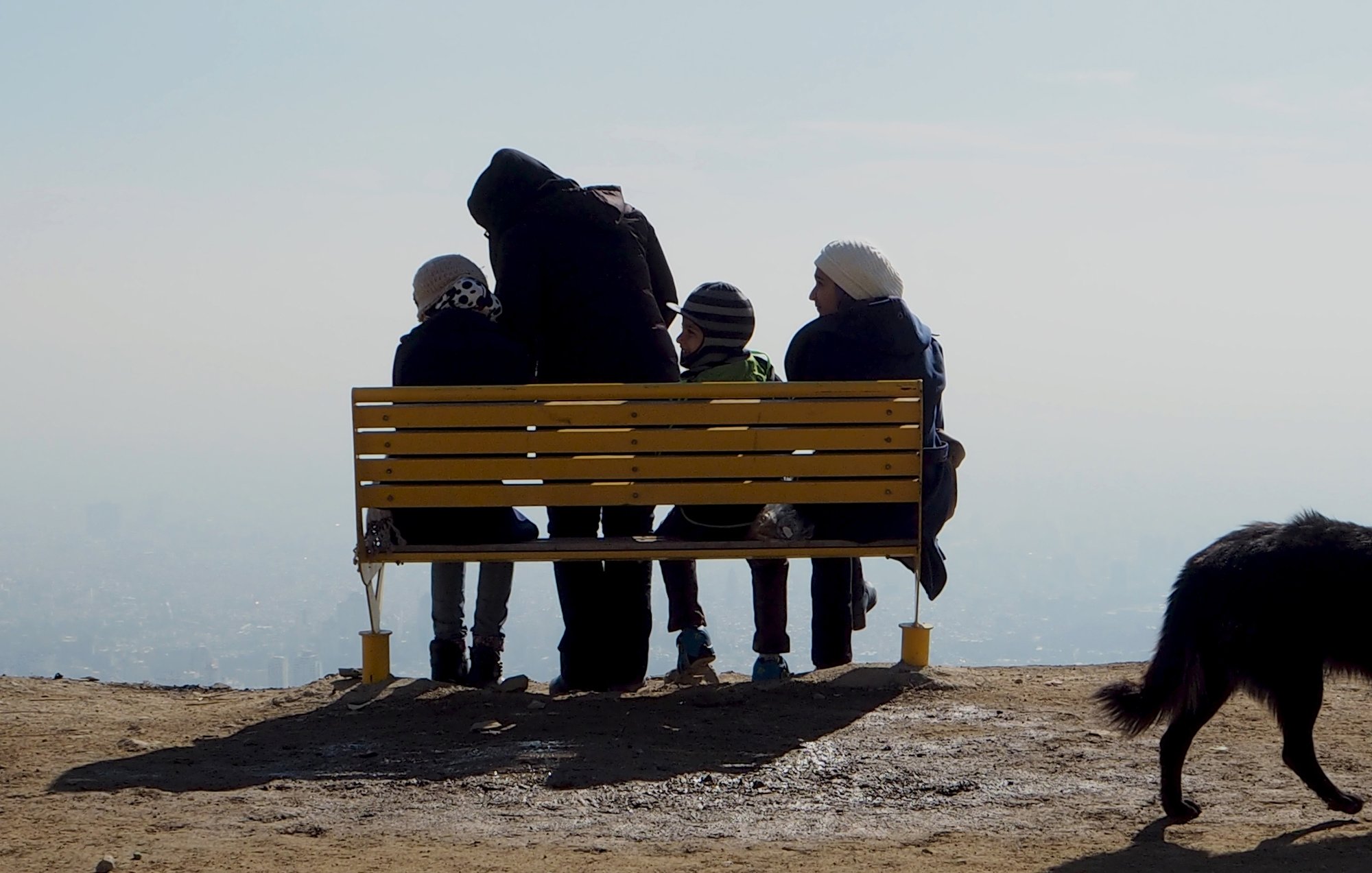در این تصویر، تعدادی از افراد در حال نشستن بر روی یک نیمکت زرد رنگ هستند. آنها به سمت یک چشمانداز زیبا و دوردست نگاه میکنند. چهار نفر در صحنه دیده میشوند: سه نفر از آنها نشستهاند و یک نفر، که احتمالاً بزرگتر است، پشت به دوربین ایستاده و به آنها توجه دارد. هوا صاف به نظر میرسد و درای پسزمینهای مهآلود است. همچنین یک سگ سیاه رنگ نیز در گوشه تصویر دیده میشود. حس کلی تصویر آرامش و دوستی را منتقل میکند.