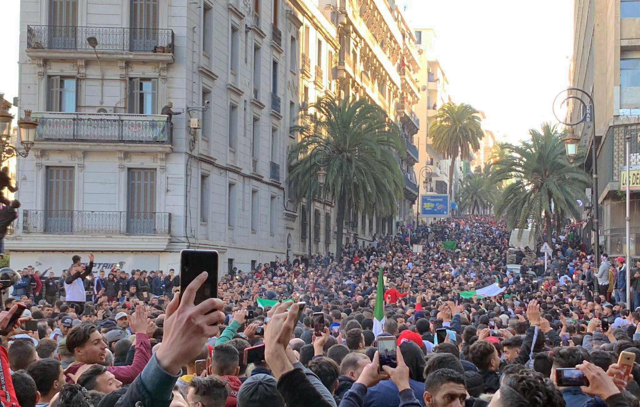 این تصویر نمایی از یک اجتماعات بزرگ در خیابانی شلوغ را نشان میدهد. مردم زیادی در حال تظاهرات یا جمع شدن هستند و در میان آنها تعدادی پرچم به چشم میخورد. درختان بلند نخل در دو طرف خیابان دیده میشوند و ساختمانهای مرتفع با بالکنها نیز در پسزمینه قرار دارند. مردم با entusiasmo در حال برنداشتن تلفنهای همراه خود و ثبت این لحظه هستند. جو عمومی تصویری از شور و شوق و همبستگی را القا میکند.