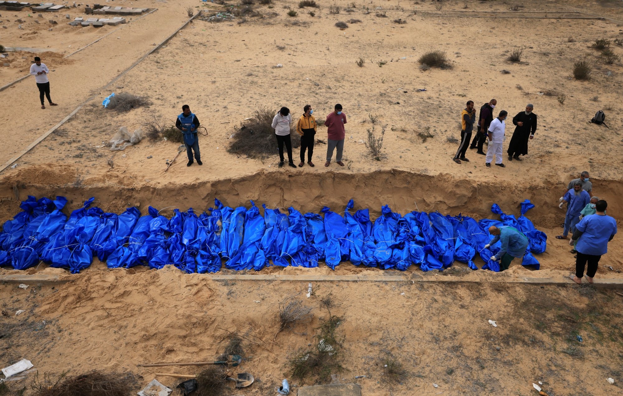 تصویر، گودالی در بیابان با تعدادی کیسه آبی رنگ و افرادی در حال مشاهده یا کار در اطراف آن است.