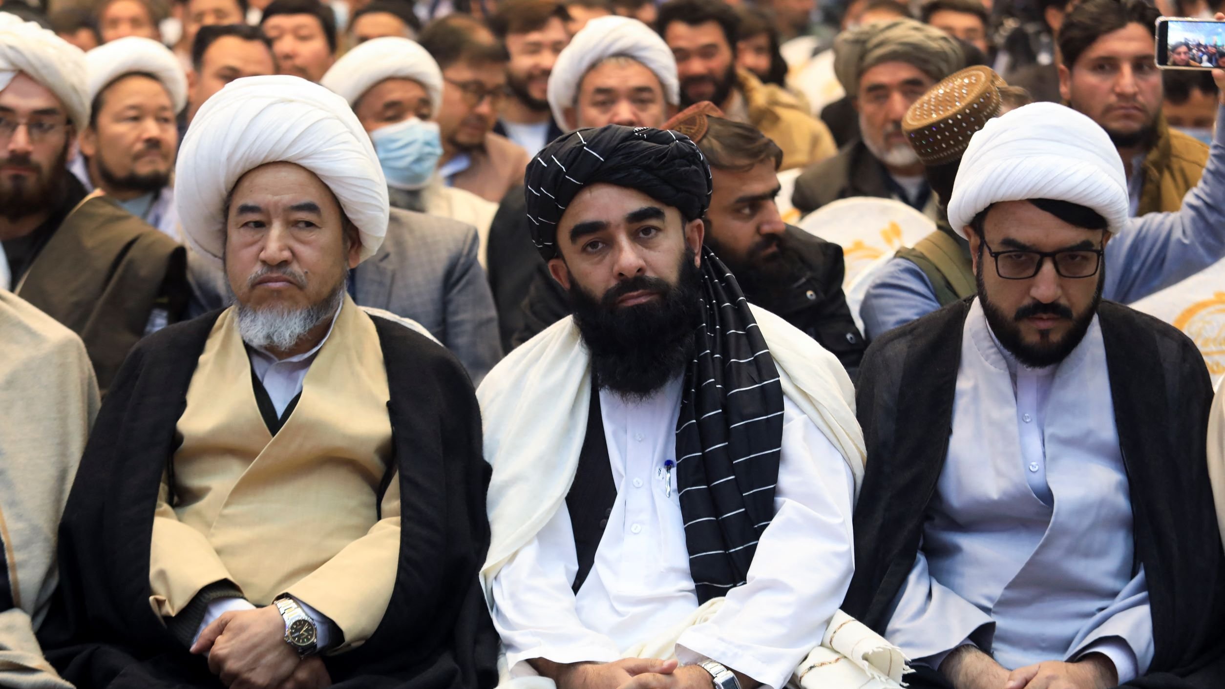 نگاهی به تصویر، گروهی از مردان را میبینیم که در یک مراسم نشستهاند. در این تصویر، سه نفر در جبهه جلو مشخصتر هستند. آنها لباسهای سنتی خود را پوشیدهاند و ظاهر آنها نشاندهنده تعلقات مذهبی و فرهنگیشان است. برخی از حضار در پسزمینه نیز قابل مشاهده هستند و به نظر میرسد این جمع در حال شرکت در یک برنامه خاص یا مراسم مذهبی هستند. فضای کلی عکس جدی و رسمی به نظر میرسد.