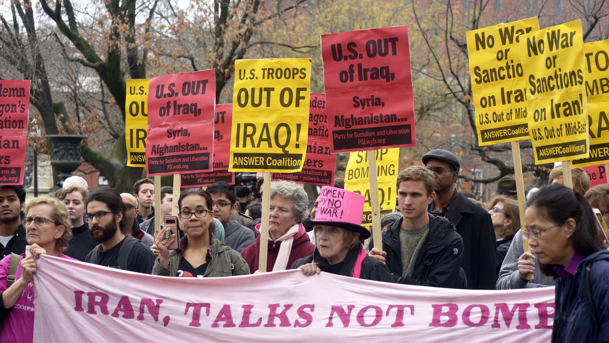 در این تصویر، گروهی از معترضان در حال برگزاری یک تظاهرات هستند. آنها پلاکاردهایی با نوشتههای مختلف در دست دارند که بر ضد دخالتهای نظامی ایالات متحده در عراق، سوریه و افغانستان و همچنین بر ضد تحریمها و جنگ تأکید میکند. همچنین، در میان جمعیت، یک بنر بزرگ با نوشته «ایران، گفتگو نه بمب» مشاهده میشود. رنگ بیشتر پلاکاردها زرد است و جمعیت نشان از تنوع دارد. فضای تظاهرات جدی و متعهدانه به نظر میرسد.
