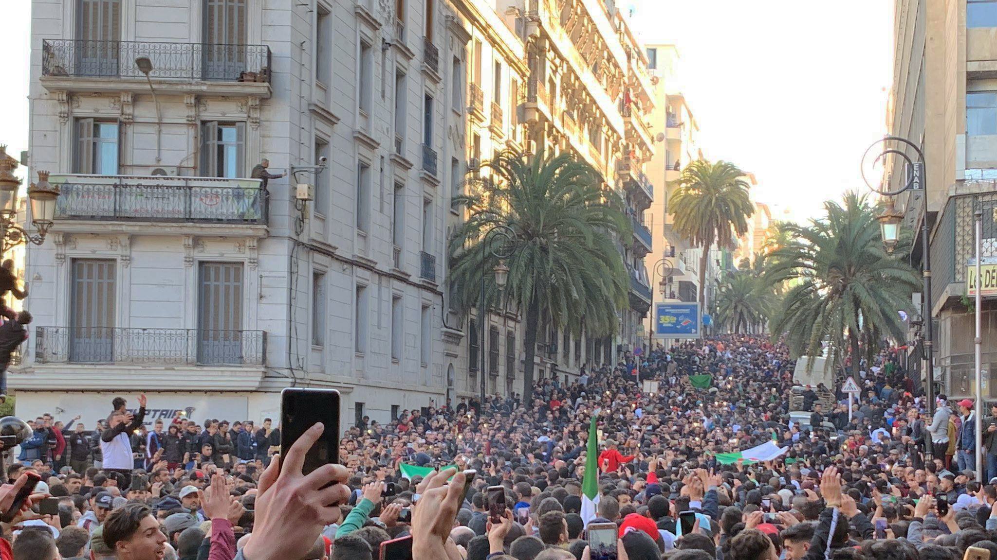 این تصویر نمایی از یک اجتماعات بزرگ در خیابانی شلوغ را نشان میدهد. مردم زیادی در حال تظاهرات یا جمع شدن هستند و در میان آنها تعدادی پرچم به چشم میخورد. درختان بلند نخل در دو طرف خیابان دیده میشوند و ساختمانهای مرتفع با بالکنها نیز در پسزمینه قرار دارند. مردم با entusiasmo در حال برنداشتن تلفنهای همراه خود و ثبت این لحظه هستند. جو عمومی تصویری از شور و شوق و همبستگی را القا میکند.