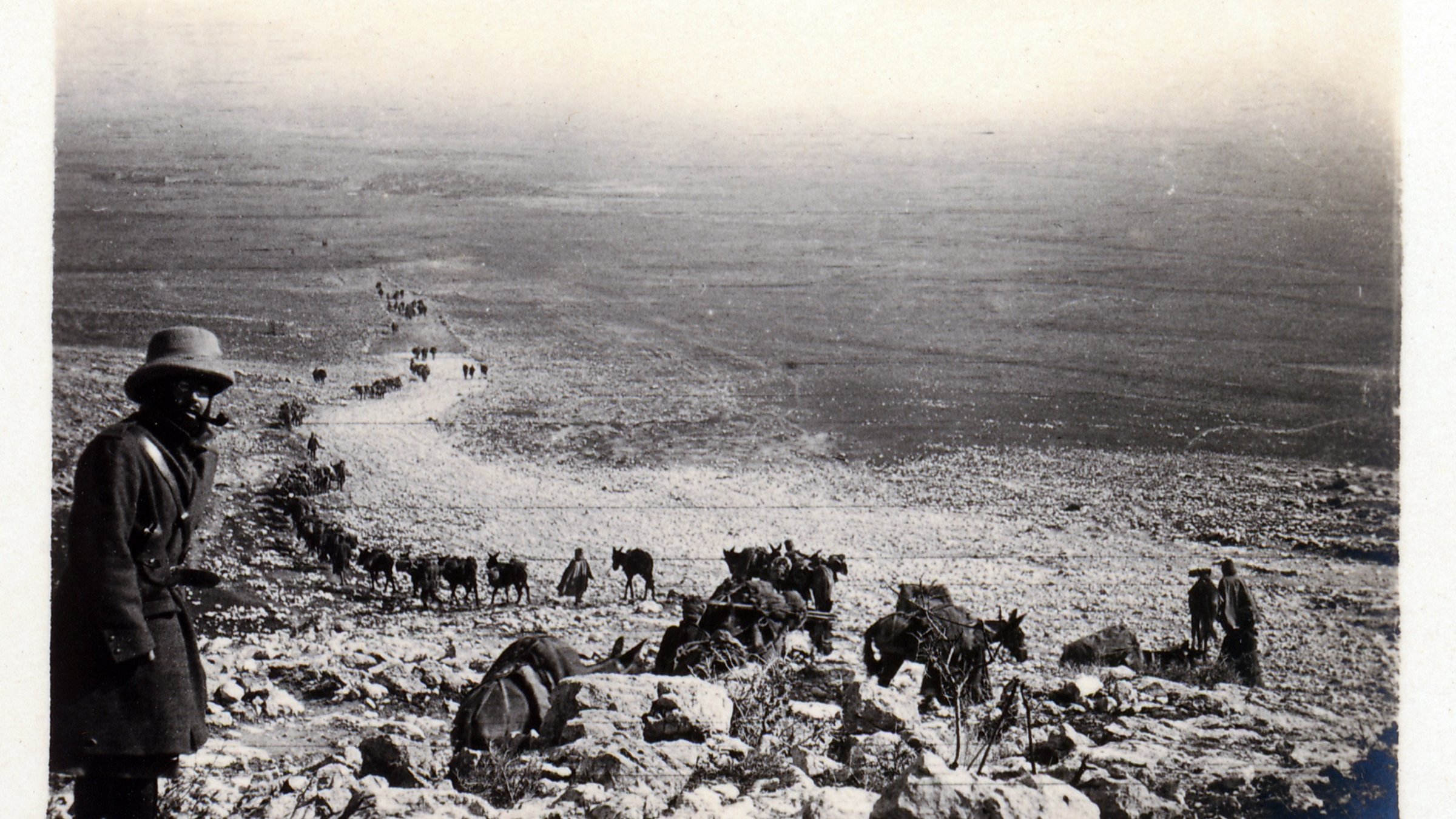 در این تصویر، مردی با کلاه و پوشاک سنتی در حال ایستادن در کنار یک شیب سنگی دیده میشود. در پسزمینه، گروهی از انسانها و حیوانات (احتمالاً خرها یا اسبها) در حال حرکت در یک مسیر سنگی به سمت پایین هستند. مناظر اطراف خشک و کوهستانی به نظر میرسد و تصویر حالتی قدیمی و سیاه و سفید دارد که به آن احساس تاریخ را القا میکند. فضای تصویر و نوع پوشش افراد نشاندهنده یک فعالیت یا سفر است.