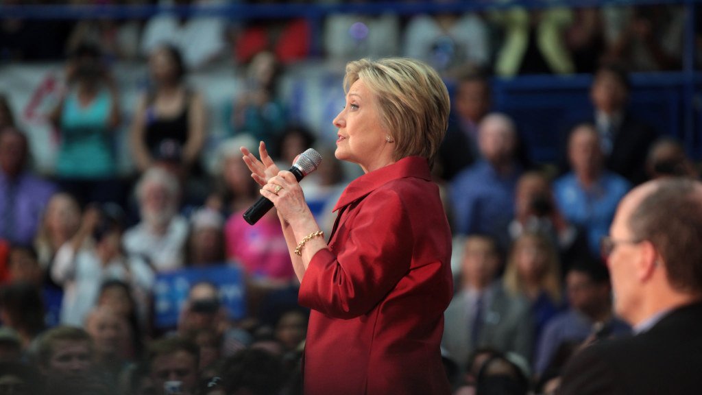 در این تصویر، فردی در حال صحبت کردن با میکروفن است. او لباسی قرمز رنگ به تن دارد و موهای بلوندش به آرامی بر روی شانههایش افتاده است. در پسزمینه، جمعیتی از افرادی که به او گوش میدهند، دیده میشوند. برخی از آنها در حال عکسبرداری یا ضبط صحبتهای او هستند. فضا به نظر میرسد که یک رویداد عمومی یا کنفرانس باشد.