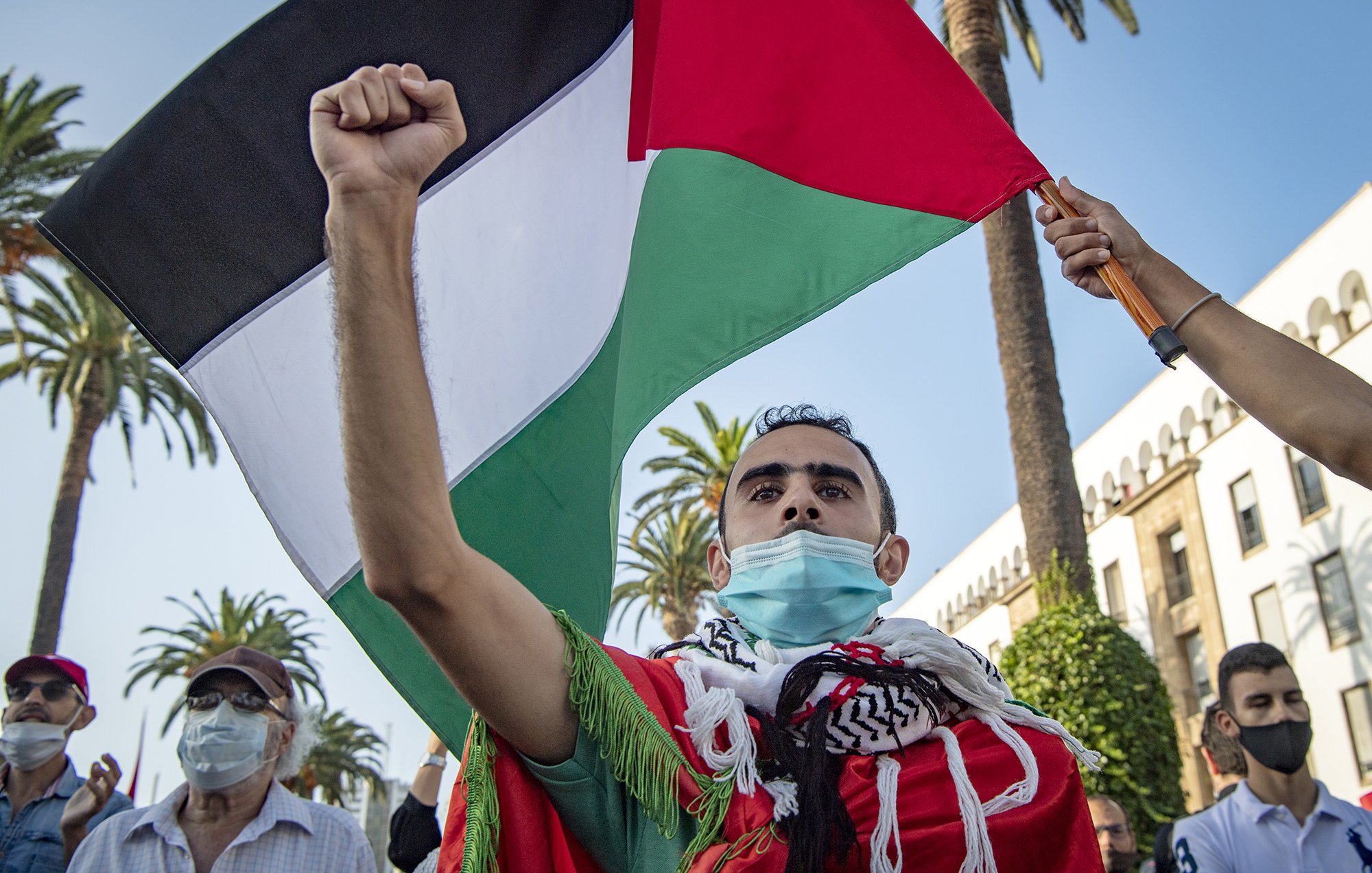 در این تصویر، مردی در حال برگزاری یک تجمع یا اعتراض است. او با افتخار پرچم فلسطین را در دست دارد و دستش را بالا برده است. چهرهاش نشاندهنده احساسات عاطفی و شور و شوق است. او همچنین ماسکی به خاطر تدابیر بهداشتی بر صورت دارد. در پسزمینه، درختان نخل دیده میشوند که به فضای گرمسیری و پرجنبوجوش این مراسم افزودهاند. افراد دیگری نیز در تصویر حاضرند که برخی از آنها ماسک زدهاند.