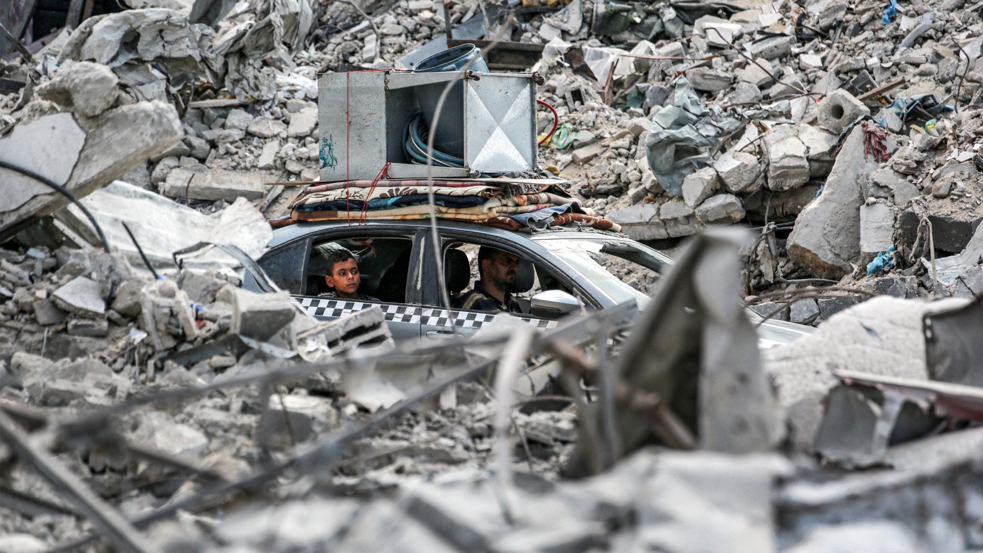 در تصویر، دو مرد درون یک خودرو در میان خرابهها نشستهاند. اطراف آنها پر از آوار و بقایای سازههای مخروبه است. برخی از أجزاء ساختمانی به وضوح دیده میشوند و وضعیتی به شدت آسیبدیده را نشان میدهند. در بالای خودرو، تجهیزات صوتی قرار گرفته است. فضای کلی تصویر حسی از ویرانی و بحران را منتقل میکند.