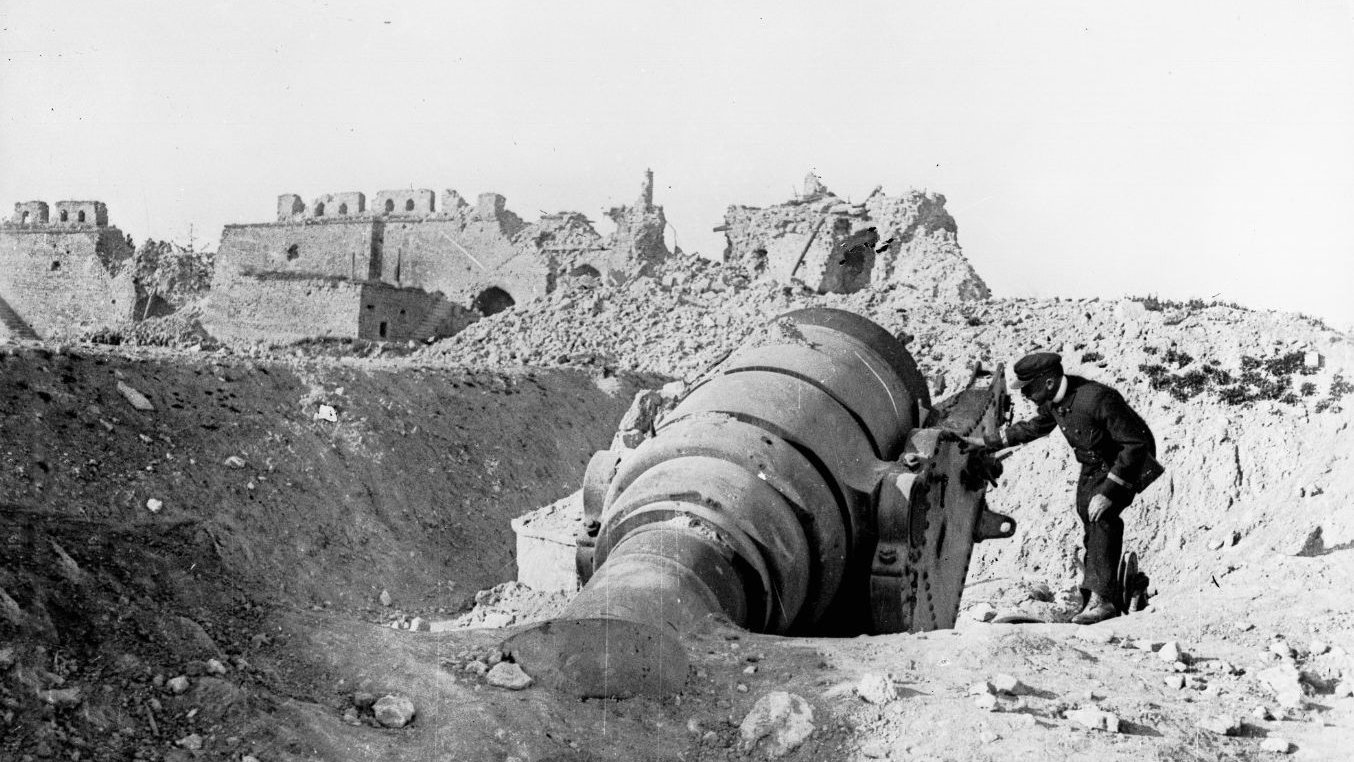 این تصویر به نظر میرسد نمایی تاریخی از یک مکان جنگی باشد. در تصویر، یک مرد به یک توپ بزرگ که به سمت زمین خوابیده است، نزدیک میشود. پسزمینه شامل ویرانههایی از ساختمانها یا قلعههای قدیمی است که گویای تخریب ناشی از جنگ یا نبردی است. همچنین، زمین اطراف آن سنگریزهها و خاکی دارد که نشاندهنده شرایط ناهموار و دشوار میباشد. به طور کلی، این تصویر حس غم و خرابی را منتقل میکند و یادآور هزینههای جنگ است.