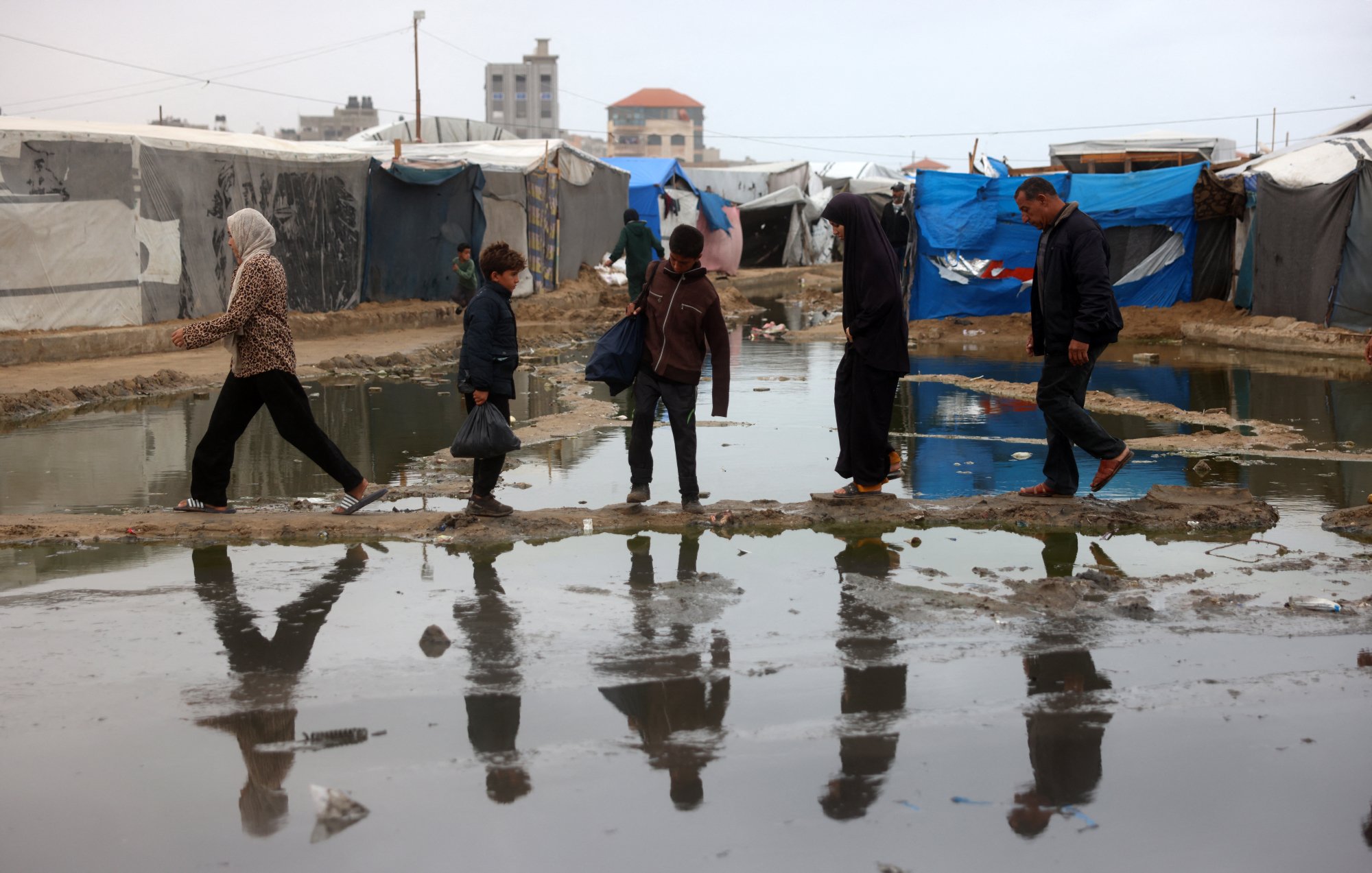 در تصویر، گروهی از مردم در یک منطقه نامناسب عبور میکنند. زیر پاهای آنها آب جمع شده و انعکاس آنها در آب دیده میشود. چادرهای موقتی در زمینه وجود دارند که به وضوح مشخص است. آسمان ابری و فضای کلی تصویر حاکی از شرایط سخت و نامساعد است. افراد در حال حرکت به سمت مقصدی مشخص هستند و لباسهای آنها نشاندهنده وضعیت اقتصادی و اجتماعیاشان است.