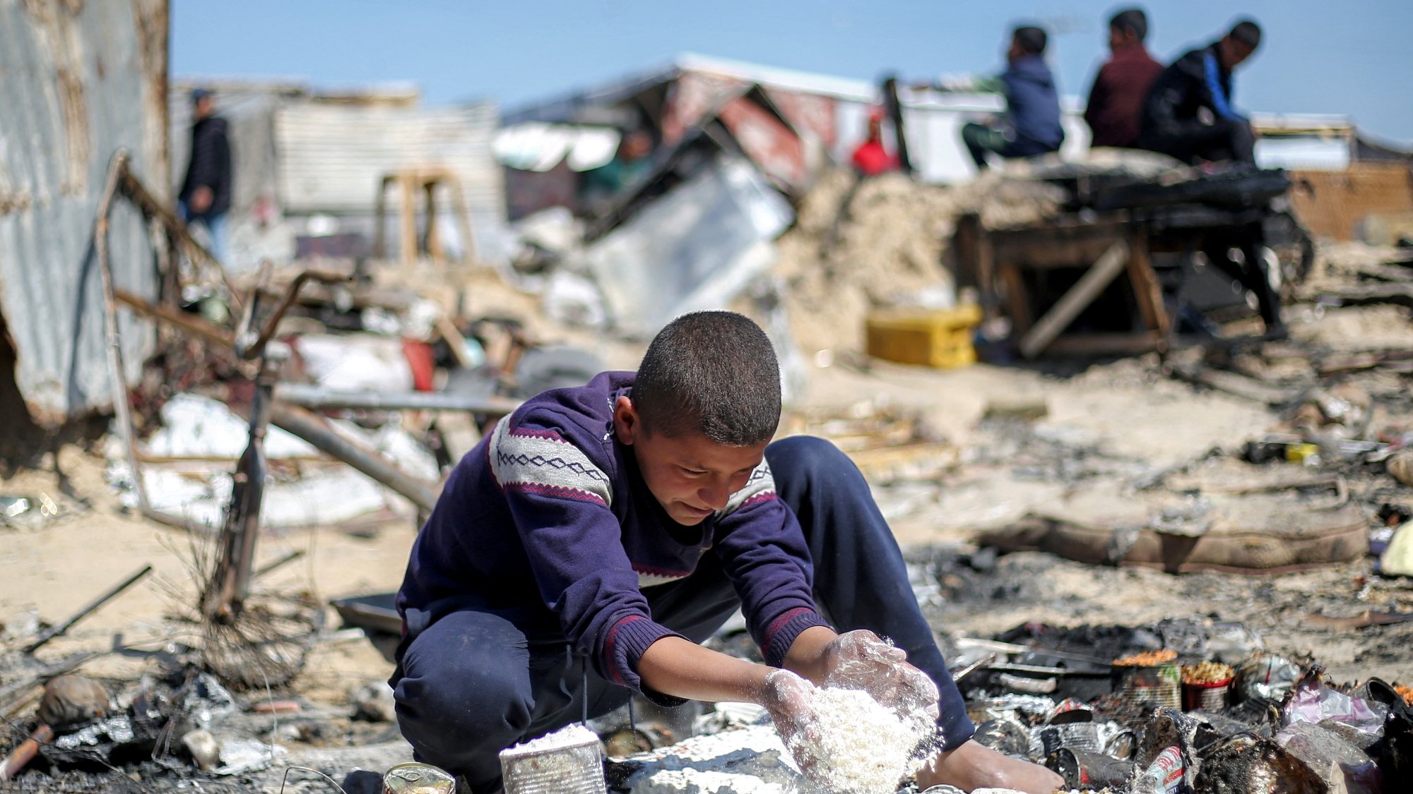 پسرکی در میان خرابهها نشسته و مشغول بازی با مواد سفید رنگ است. فضای اطراف بسیار بینظم و ویران است.